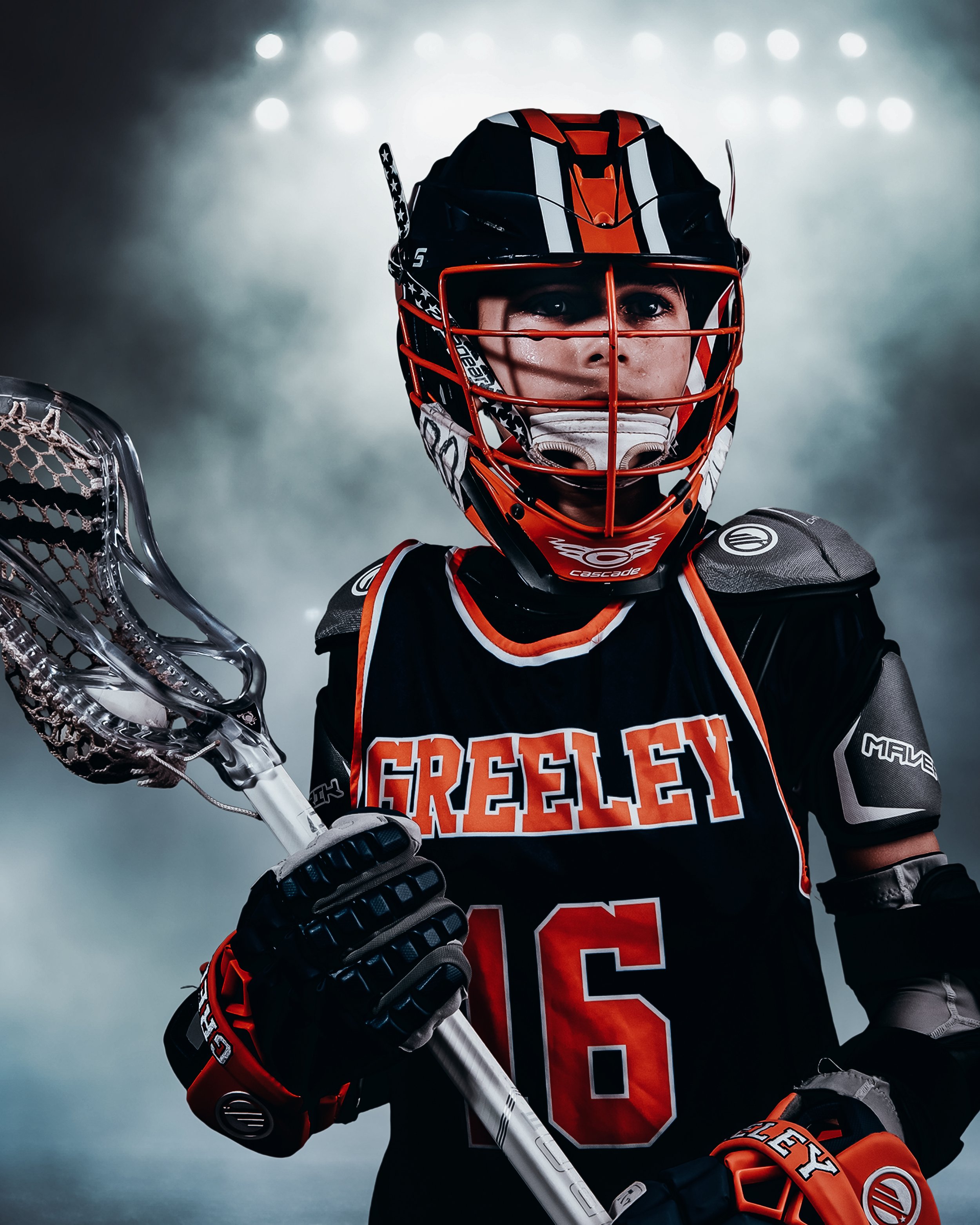 Lacrosse player in full gear holding a lacrosse stick, wearing a black and orange team jersey with 'GREELEY' and the number 16, a helmet with a cage, gloves, and shoulder pads, against a smoky background.