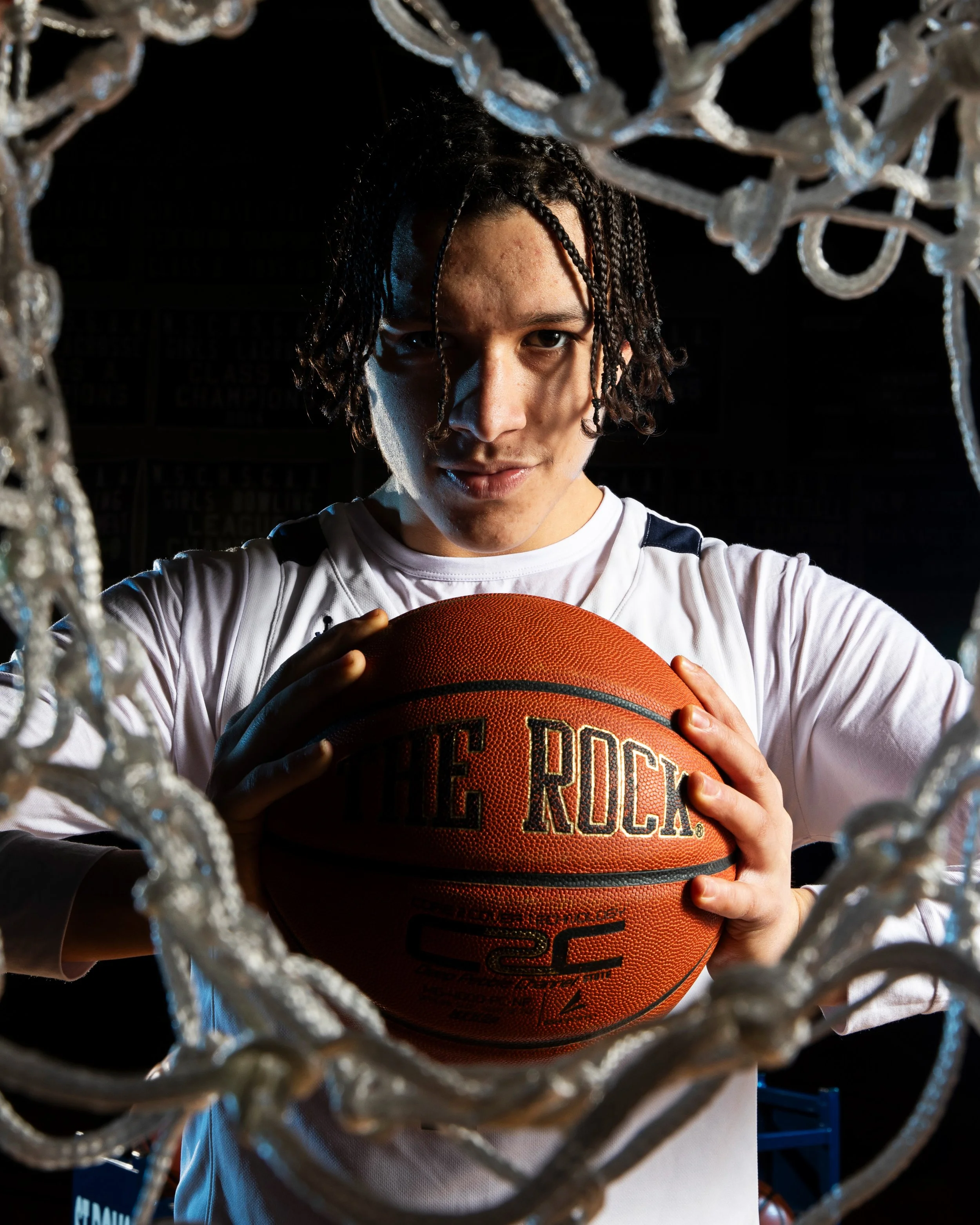 A young basketball player with braided hair, holding an orange basketball with the words 'The Rock' on it, standing behind a basketball net in a dark gymnasium.
