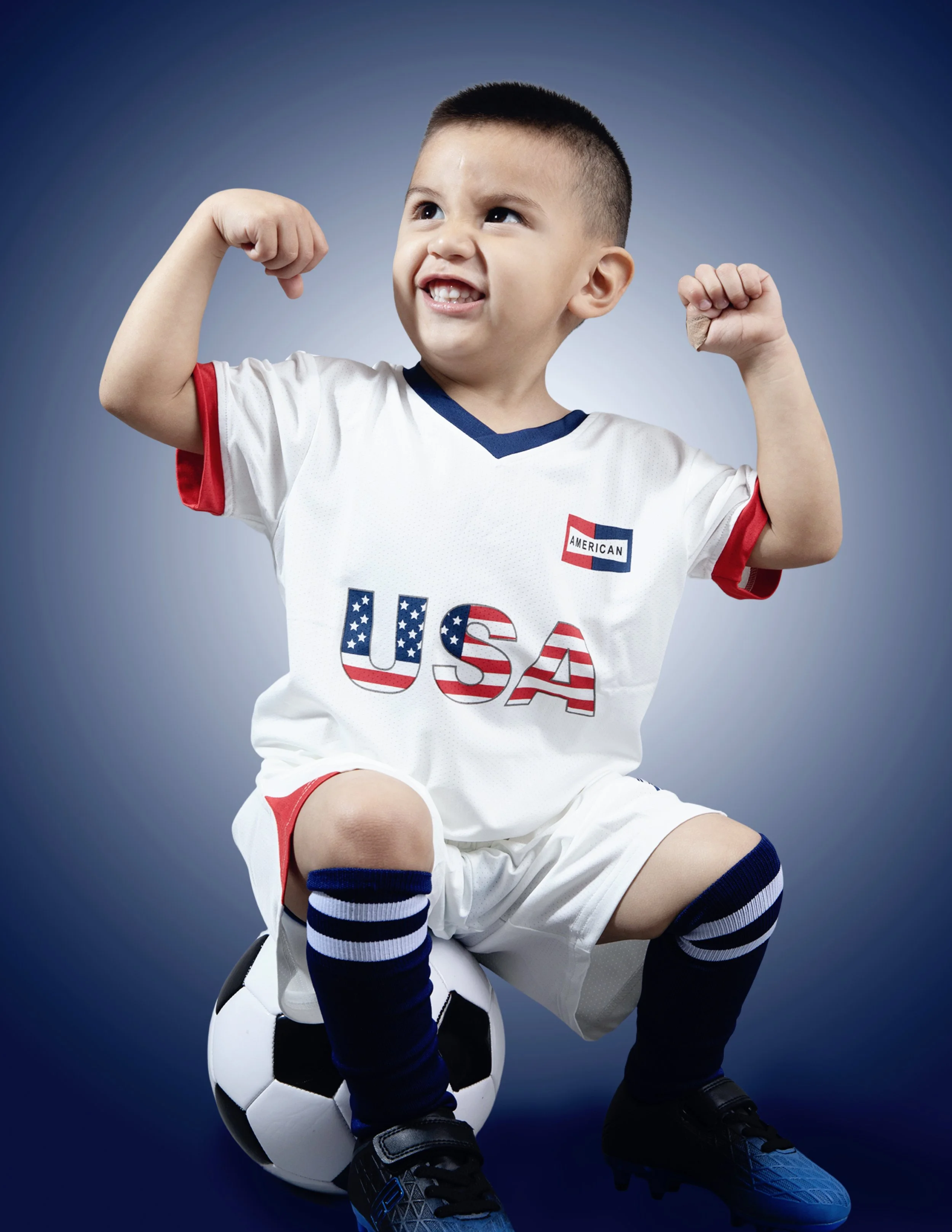 Young boy in USA soccer uniform sitting on a soccer ball, flexing his arms with a confident expression, against a plain background.