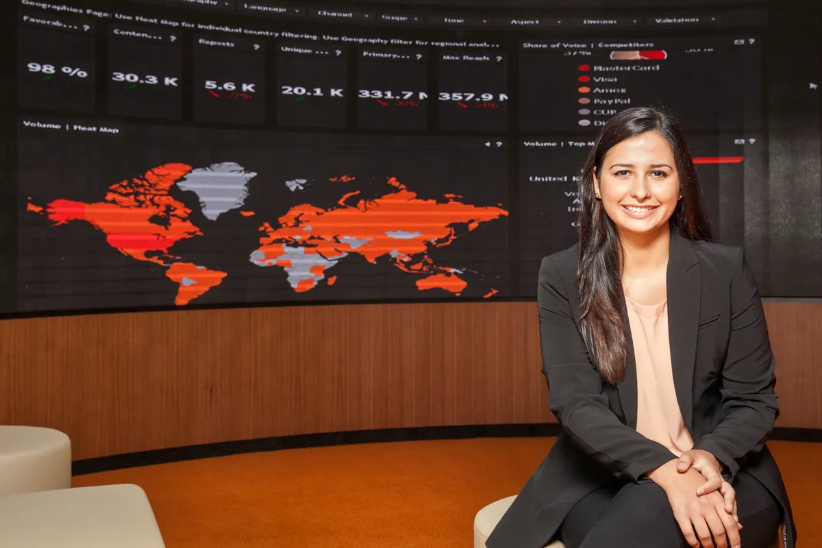 A woman sitting on a chair smiling in front of a large digital world map with data visualizations in a conference room.