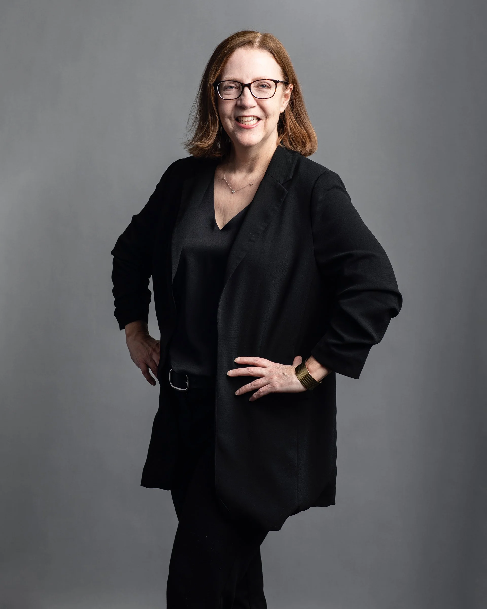 A woman with shoulder-length reddish brown hair, wearing glasses, a black blazer, and a black top, standing with one hand on her hip against a plain gray background.