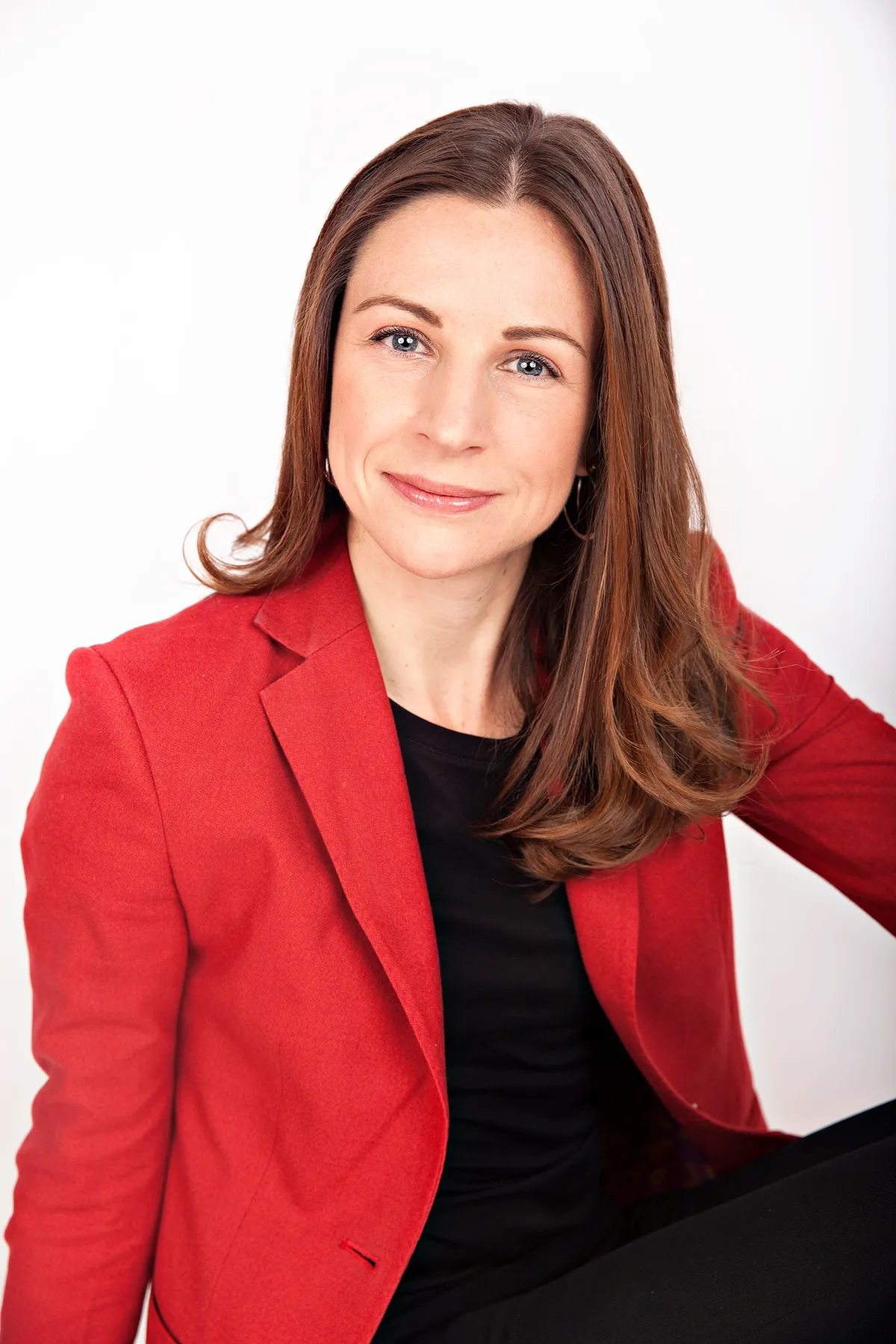 A woman with shoulder-length brown hair, wearing a red blazer and black top, smiling and sitting against a plain white background.