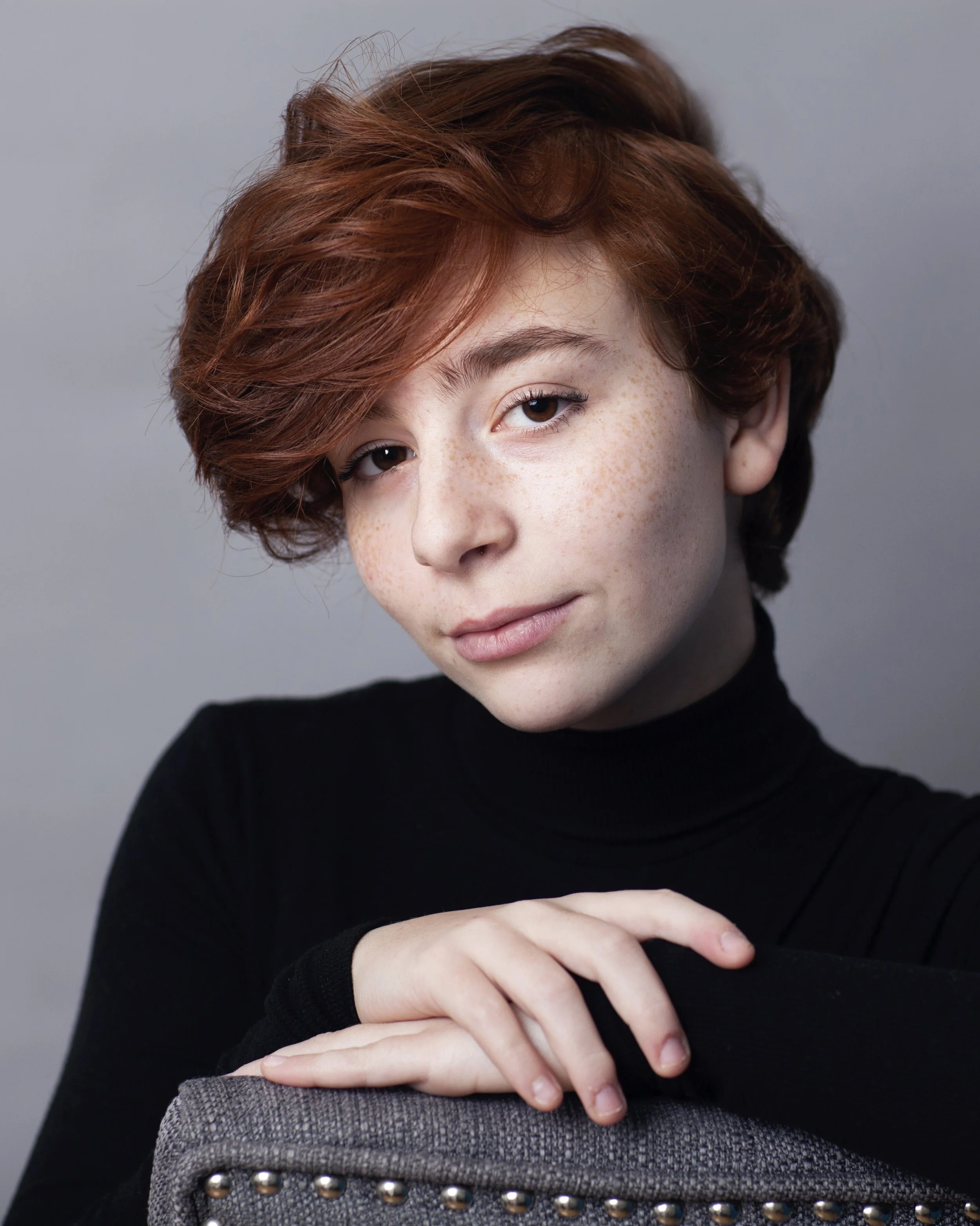 A young woman with short red hair, a black turtleneck, and freckles resting her arm on a gray upholstered chair with metal studs, looking at the camera.