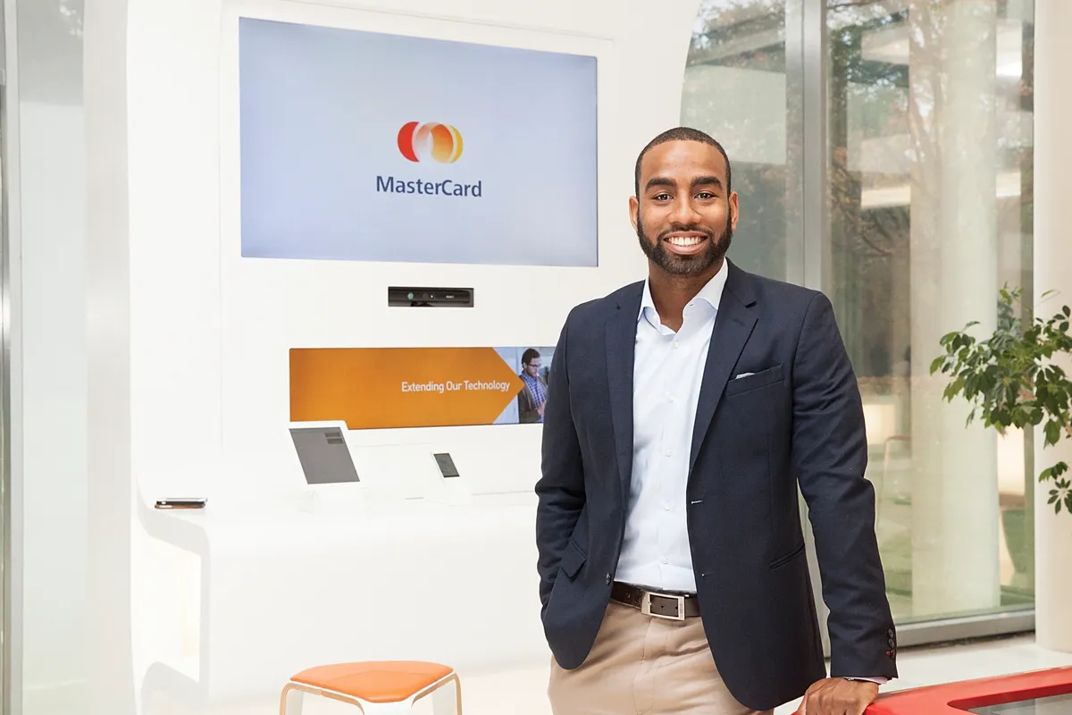 A man in business attire smiling in front of a MasterCard kiosk in a modern office or retail space.