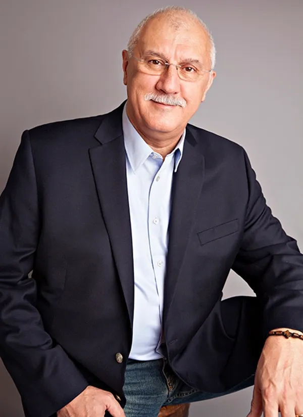 A senior man with glasses and a mustache wearing a dark blazer and light blue shirt, sitting against a neutral background.