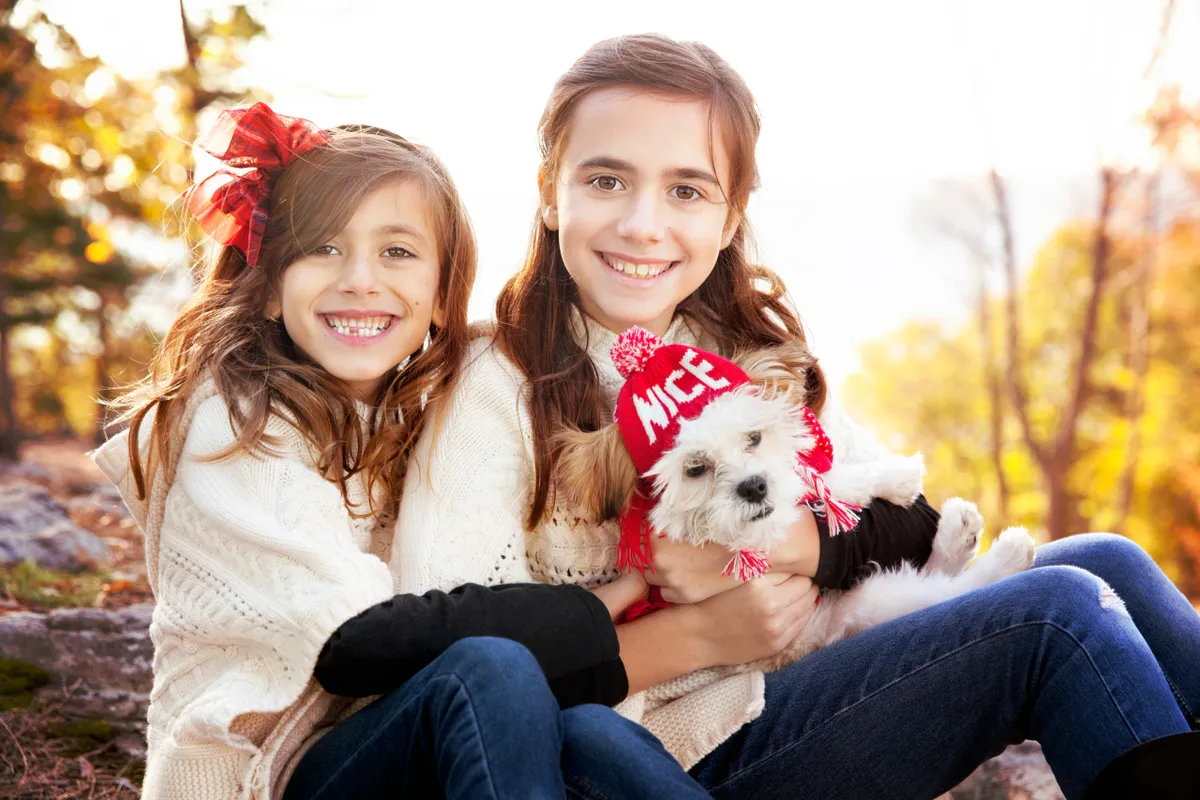 Two young girls sitting outdoors in autumn, smiling and holding a small white dog wearing a red hat and scarf. The background features trees with fall foliage.