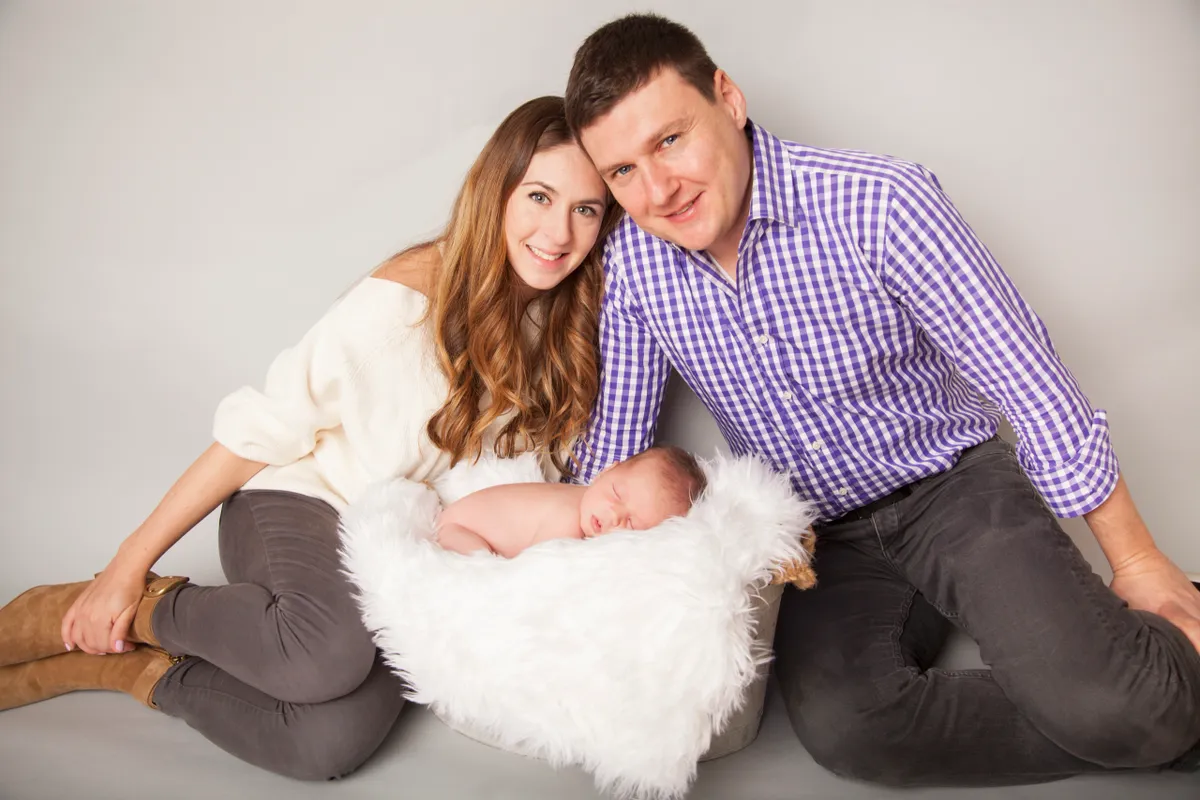 A family of three, including a mother, father, and newborn baby, posing together on a gray background. The mother and father are kneeling, with the baby sleeping on a white fluffy blanket between them. The mother has long, wavy brown hair and is wear
