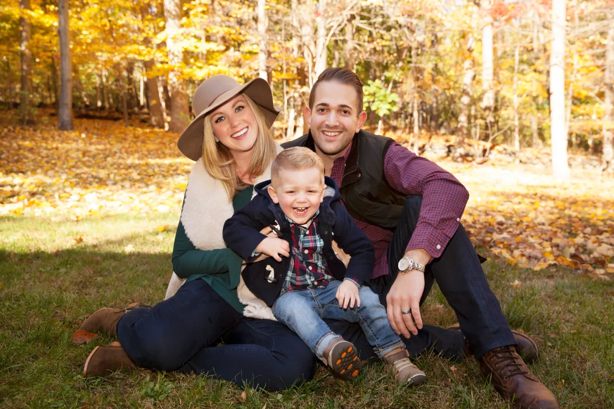 A smiling family of three sitting on the grass in a park during fall, surrounded by trees with orange, yellow, and brown leaves.