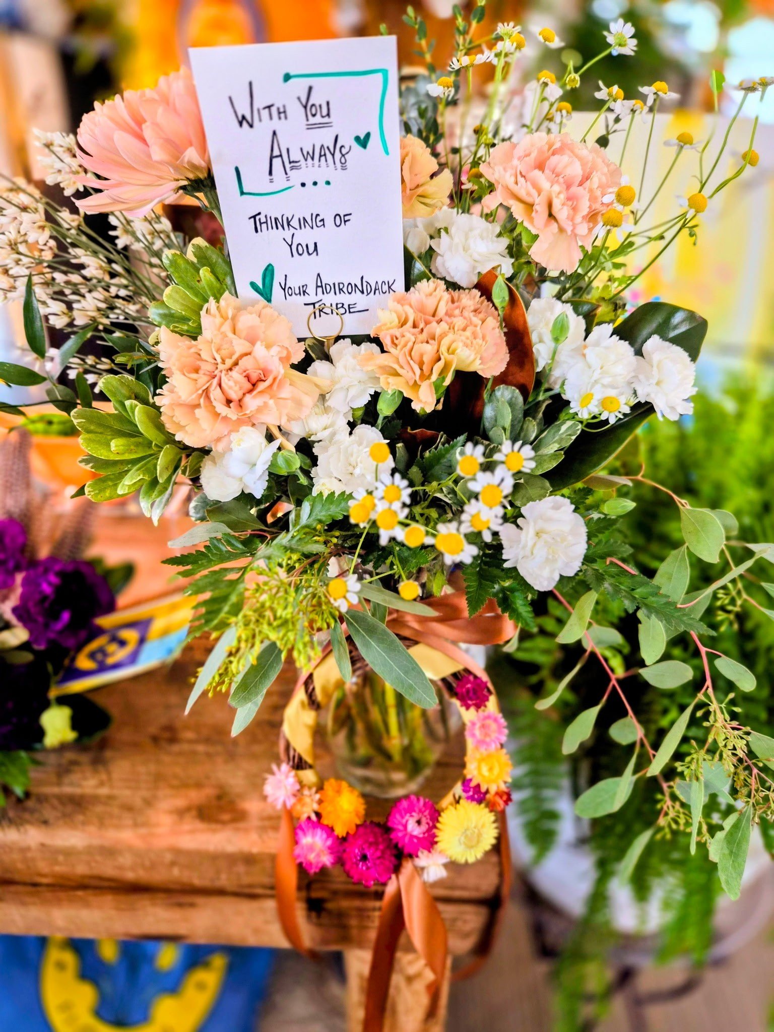 A bouquet of pink, white, yellow, and purple flowers with a handwritten note that reads, 'With You Always, Thinking of You, Your Adirondack Tribe,' is displayed in a glass vase on a wooden table, decorated with a ribbon. The background includes other floral arrangements.