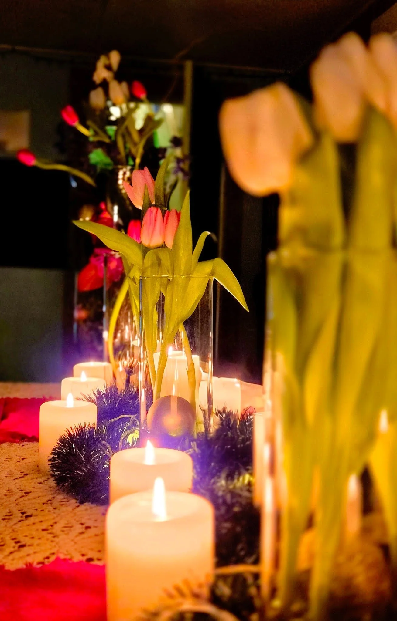 Vase with pink tulips, surrounded by lit candles, candles, black garland on table, and blurred floral arrangement in background.