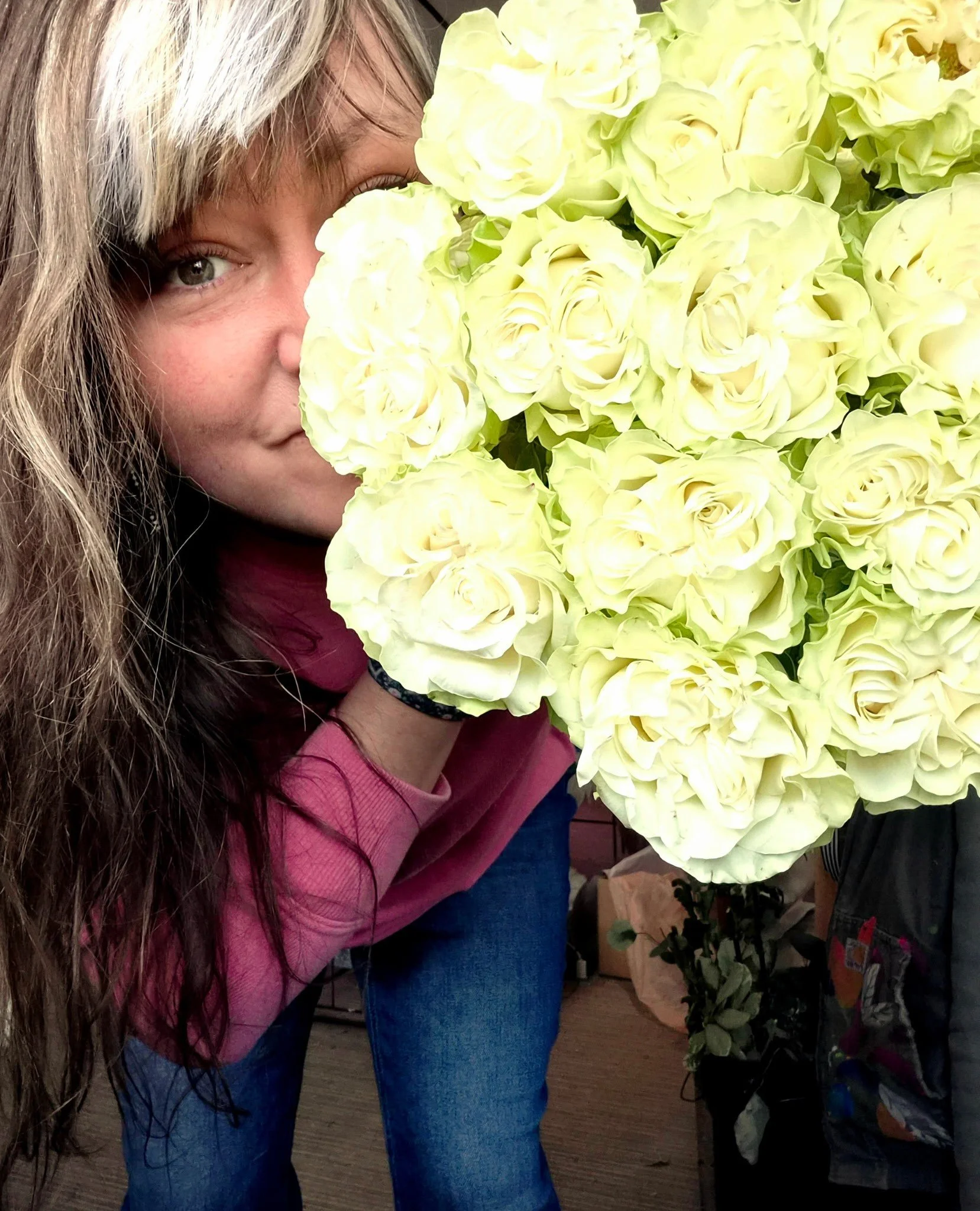 Woman with long hair and a pink sweater smelling a large bouquet of cream-colored roses.
