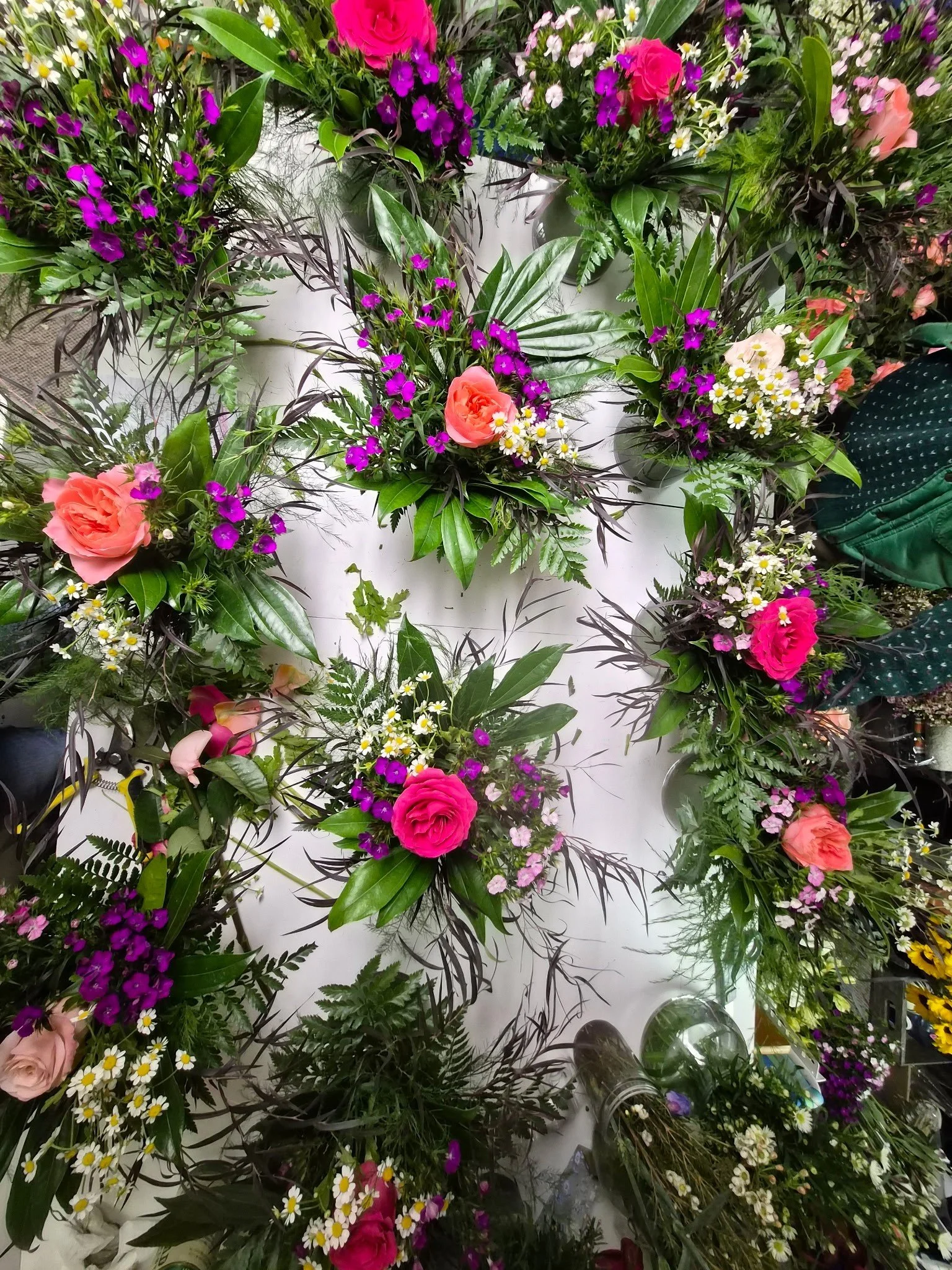 A wall decorated with numerous floral arrangements, featuring pink roses, purple and white daisies, and other green foliage.