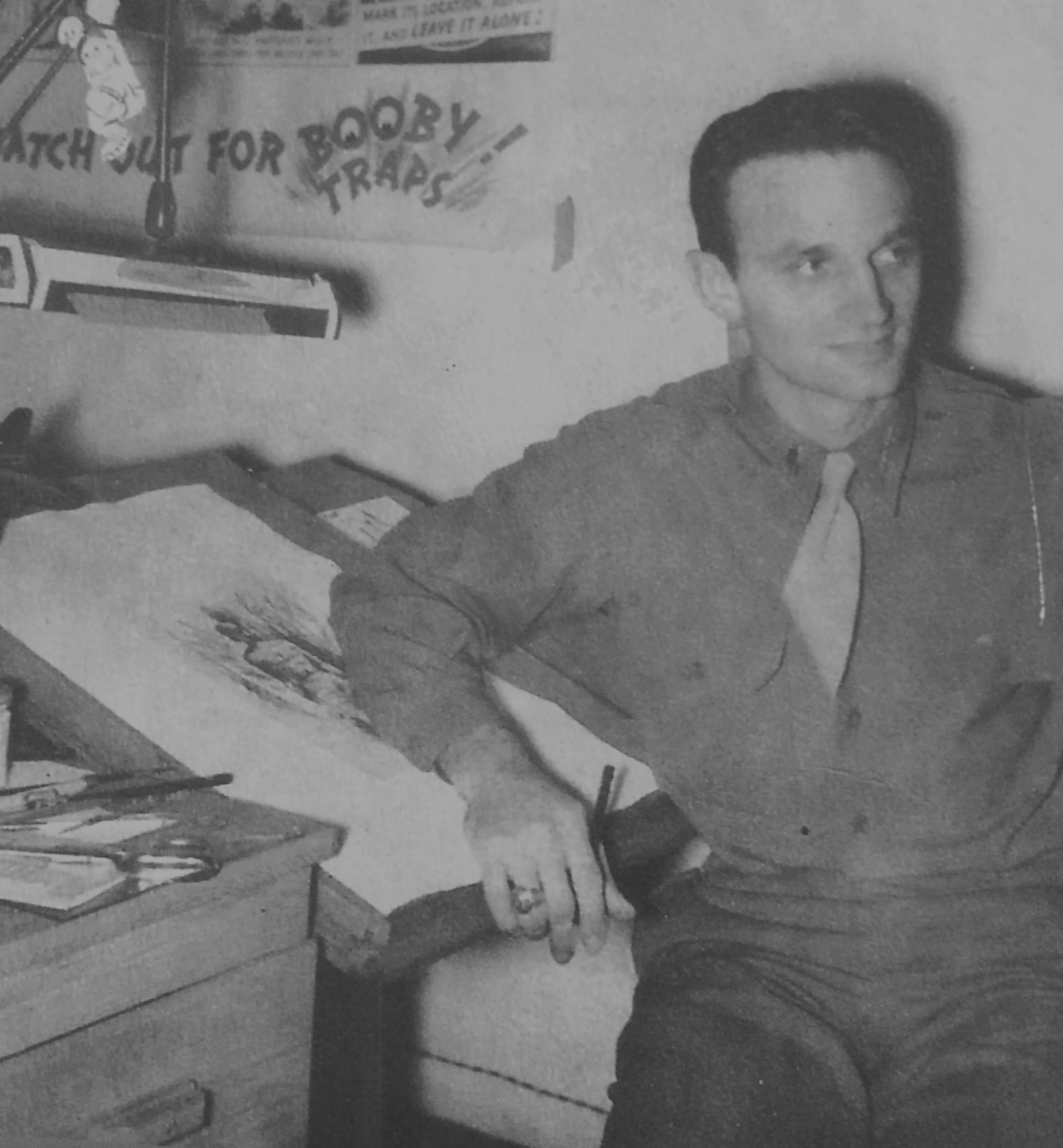 A young man with dark hair, wearing a button-up shirt and a necktie, sitting at a desk with art supplies, holding a pen, with a poster on the wall behind him reading 'Watch out for bloody traps!'