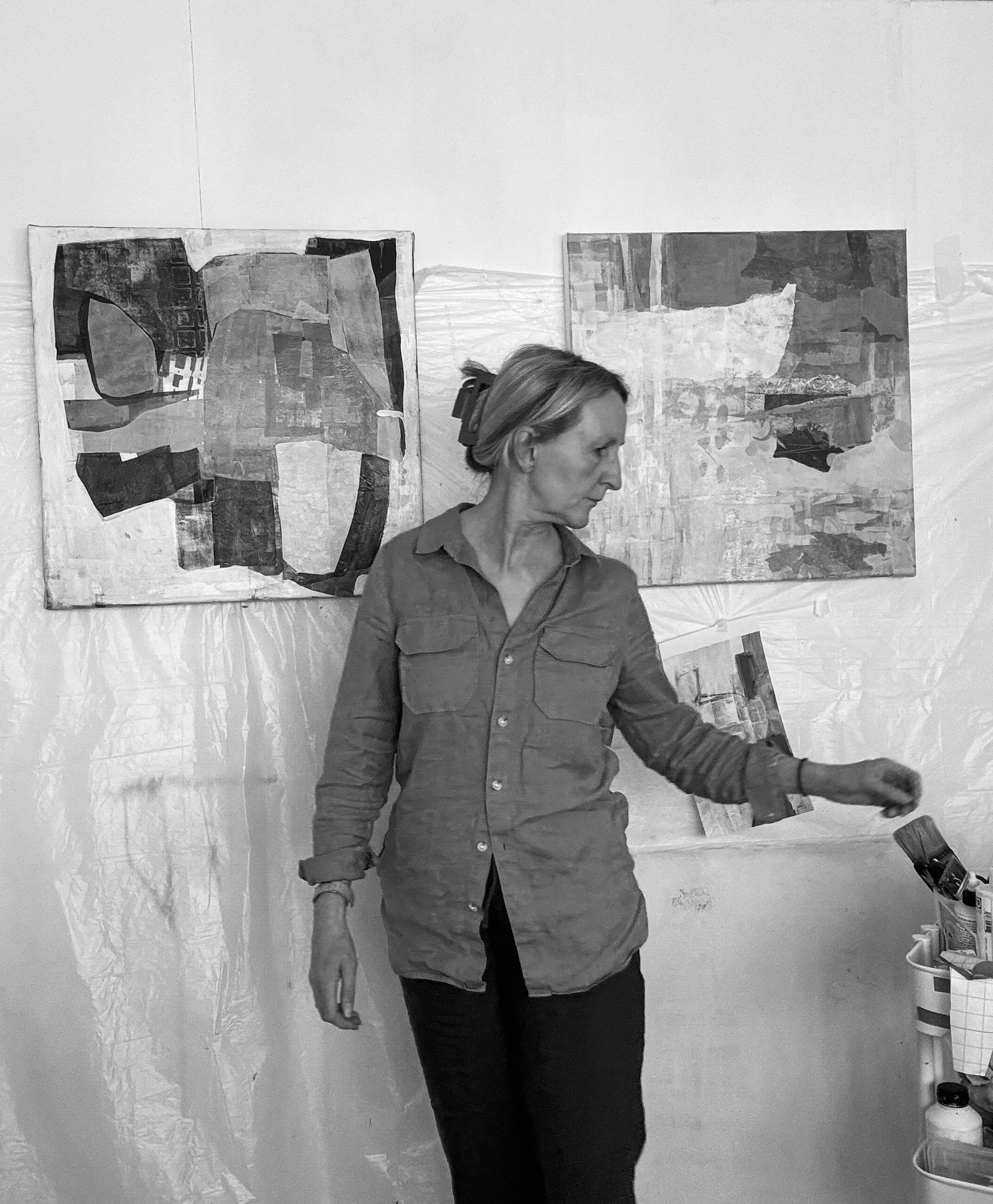 Black and white photo of Artist Angela Slack in the studio painting. She is stood in front of two all mounted canvases