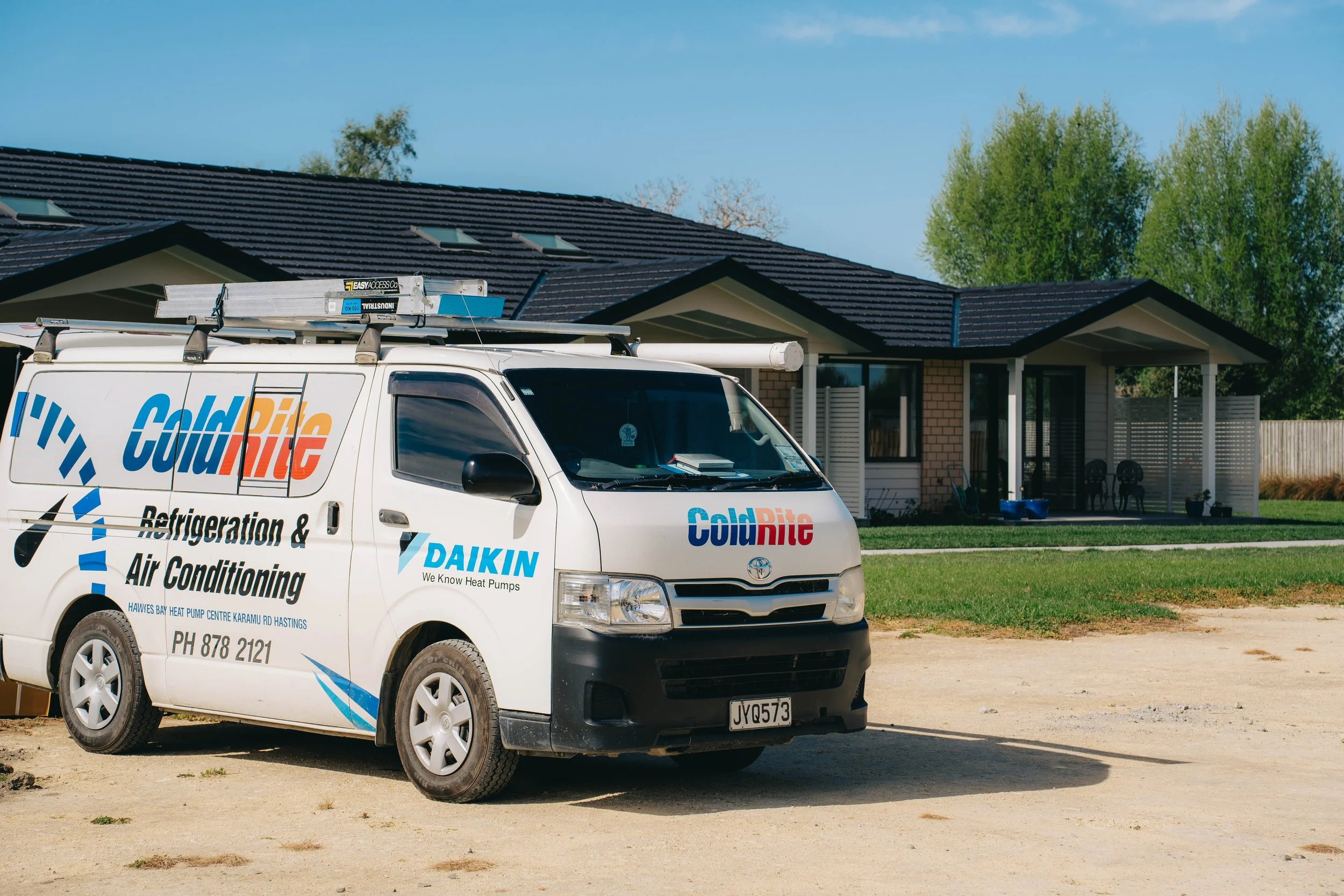 A white service van with branding for Cold Rite Refrigeration and Air Conditioning parked on a dirt driveway in front of a residential house with a dark tiled roof. The van is equipped with a ladder on the roof and shows the contact information for the business. The house has a porch and is surrounded by green trees.