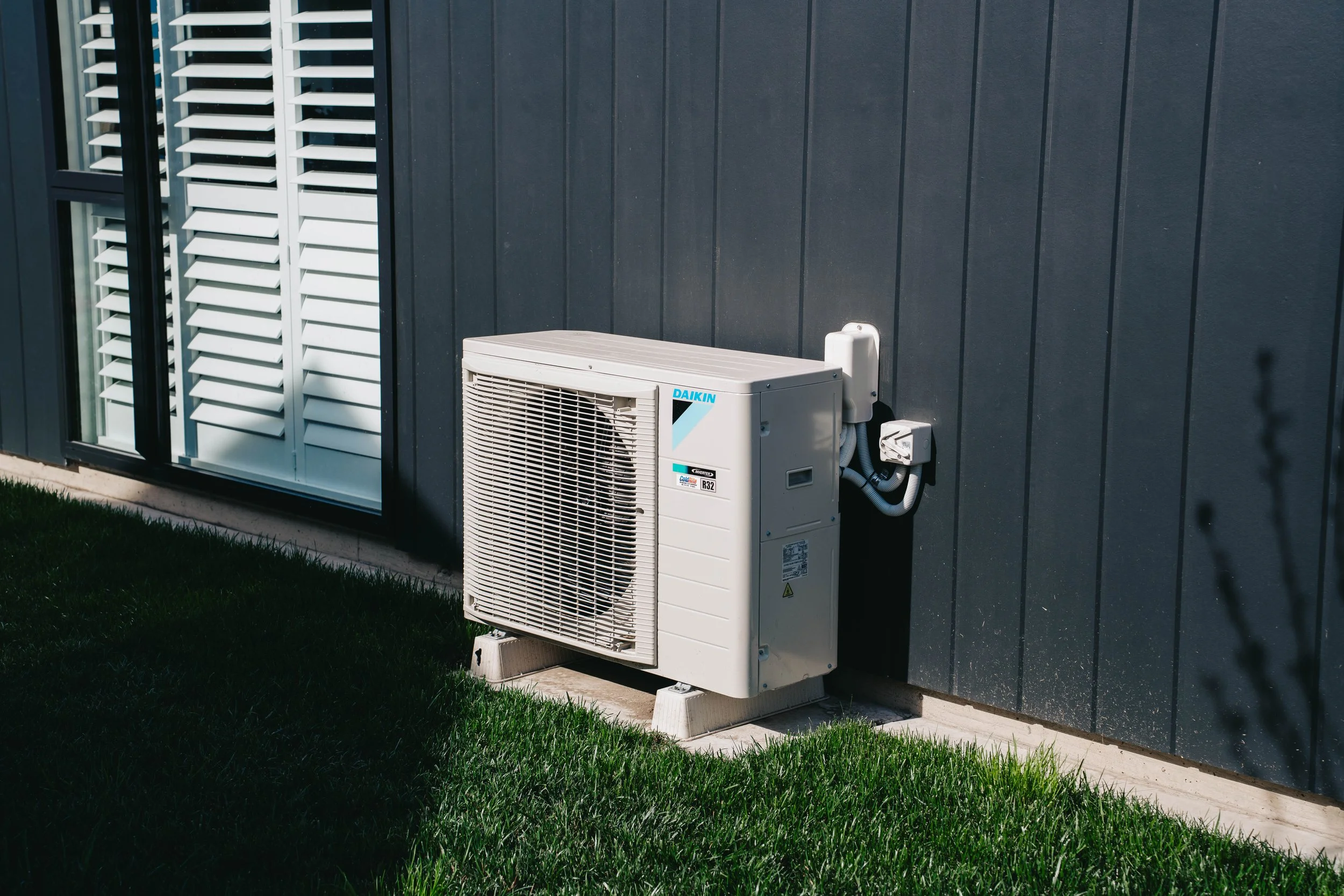 Exterior view of a dark-colored house with an air conditioning unit installed outside. The unit is mounted on a platform near a wall with vertical siding. There is a large window with white shutters to the left of the unit, and a patch of green grass in the foreground.