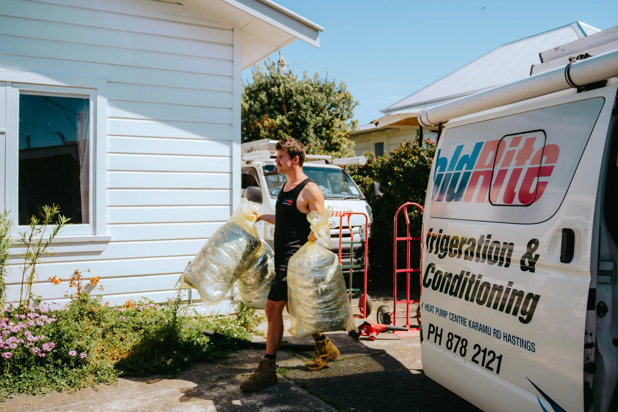 A man in work boots and a sleeveless shirt carrying large plastic bags of leaves or yard debris on a residential driveway, near a white house with a garden and a yard equipment truck with company signage.