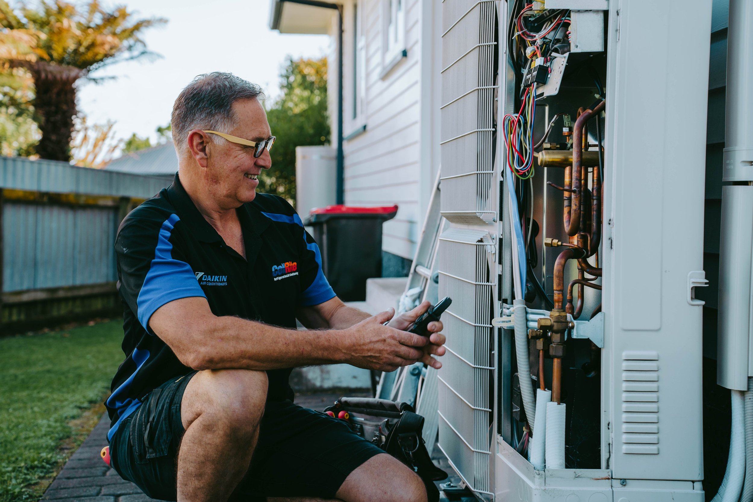 A technician wearing a black and blue uniform kneels outdoors next to an open air conditioning unit, performing maintenance using a multimeter.