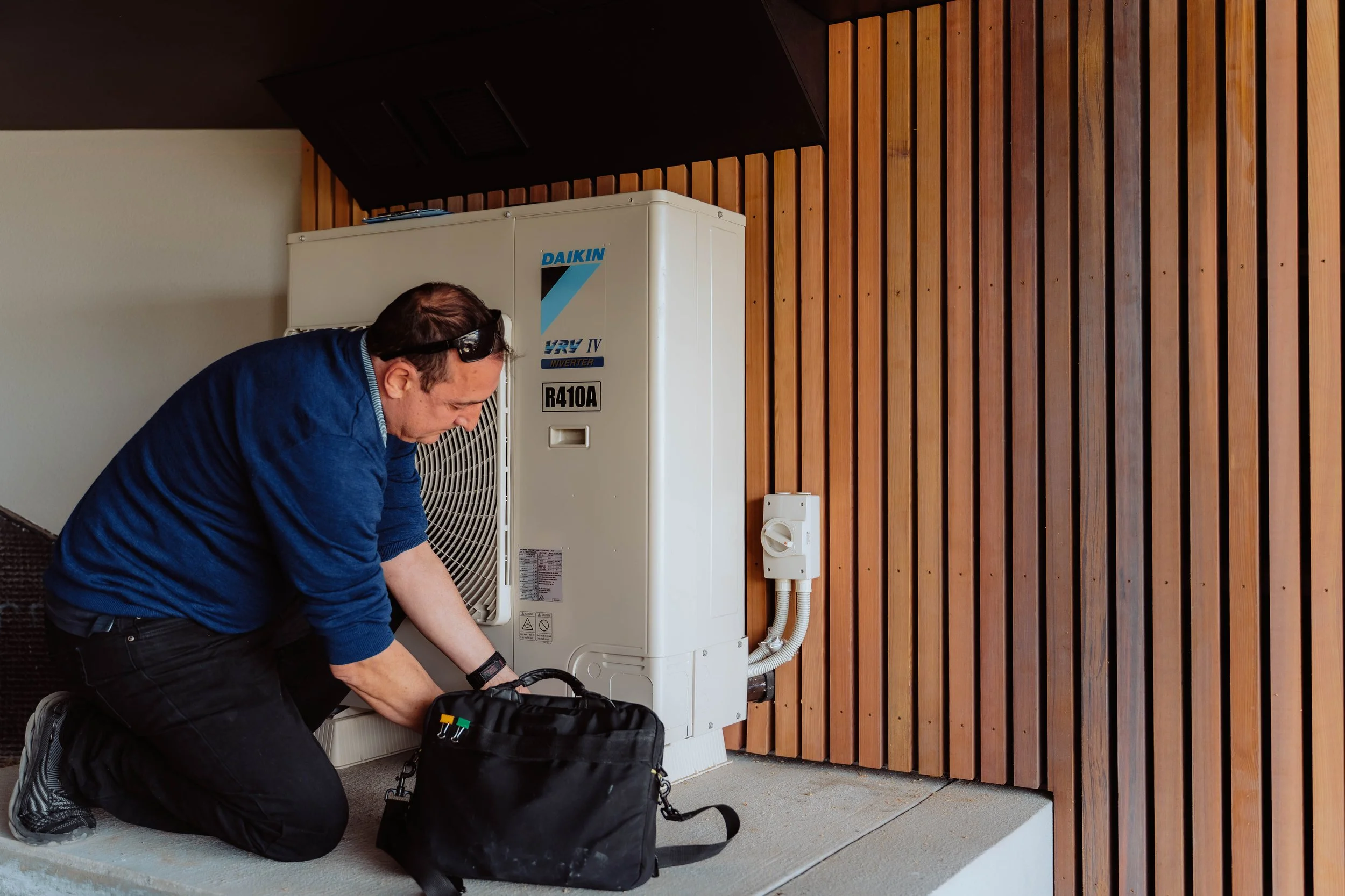 A technician working on an outdoor HVAC unit next to a wooden wall, wearing a blue jacket and safety glasses, with a toolkit open on the ground.