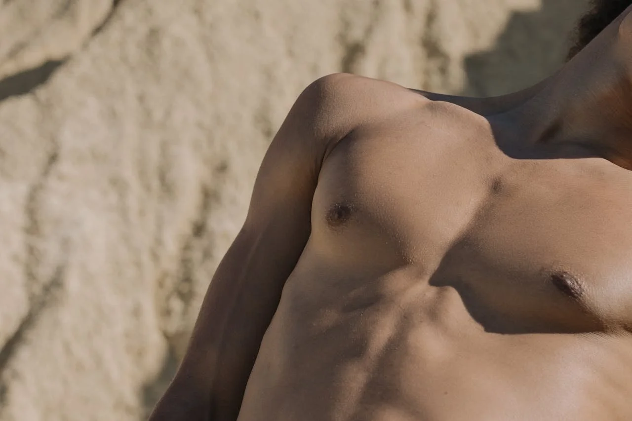 Close-up of a shirtless person's upper body, focusing on the shoulder, chest, and abdomen, with a stone wall in the background.