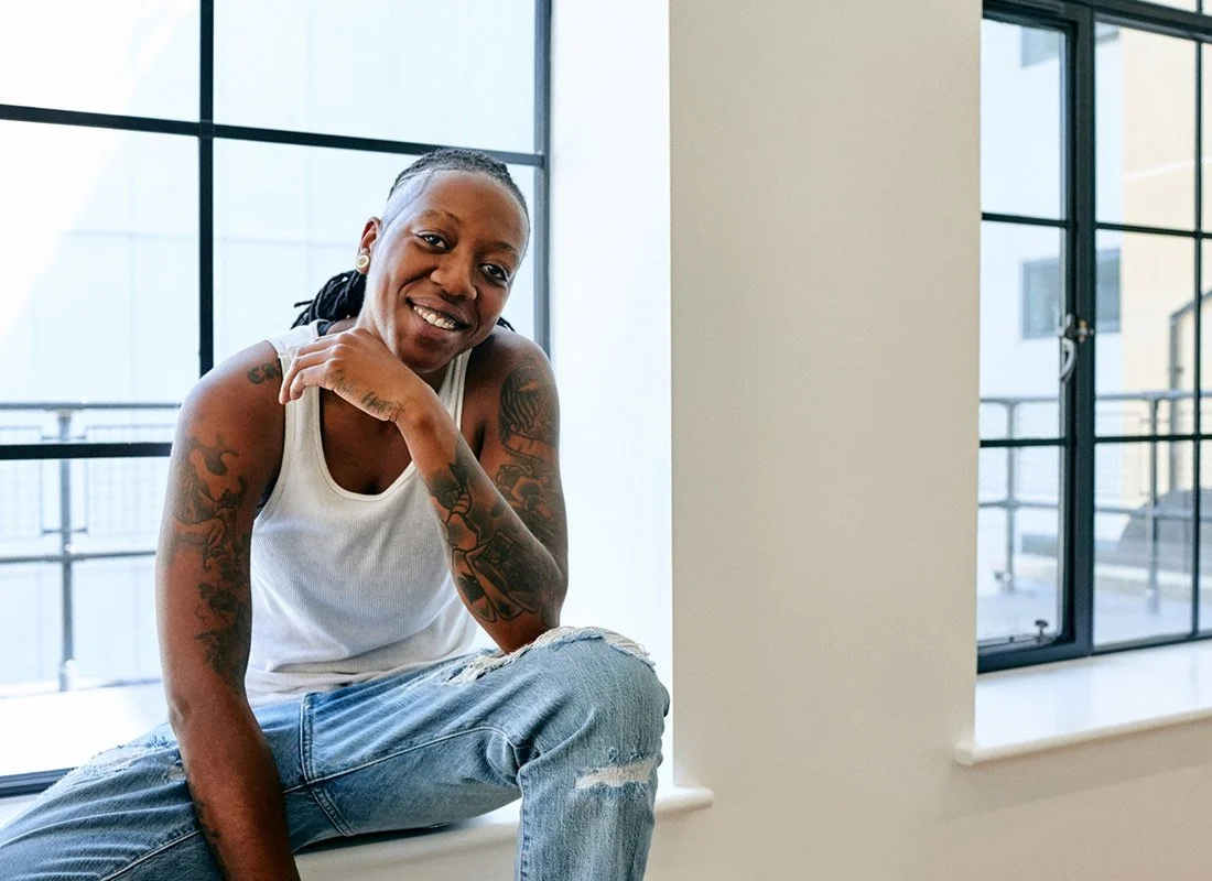A smiling woman with tattoos on her arms, wearing a white sleeveless top and ripped jeans, sitting on a window sill in a bright, modern room with large windows.