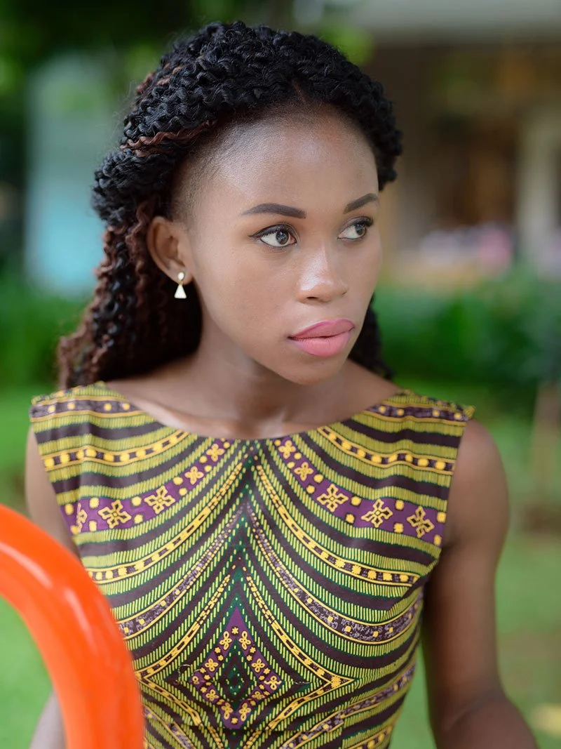 A young woman with dark curly hair styled in twists, wearing a colorful traditional African print dress with geometric patterns in yellow, green, and purple, standing outdoors with a blurred green background.