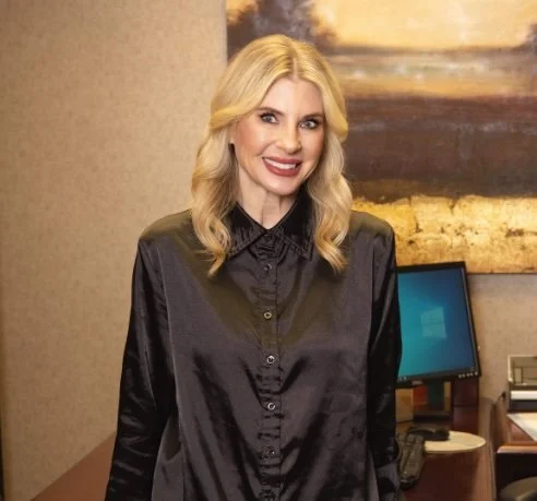 A woman with blonde hair and a black satin blouse smiling indoors, standing in front of a computer monitor and abstract art on the wall.