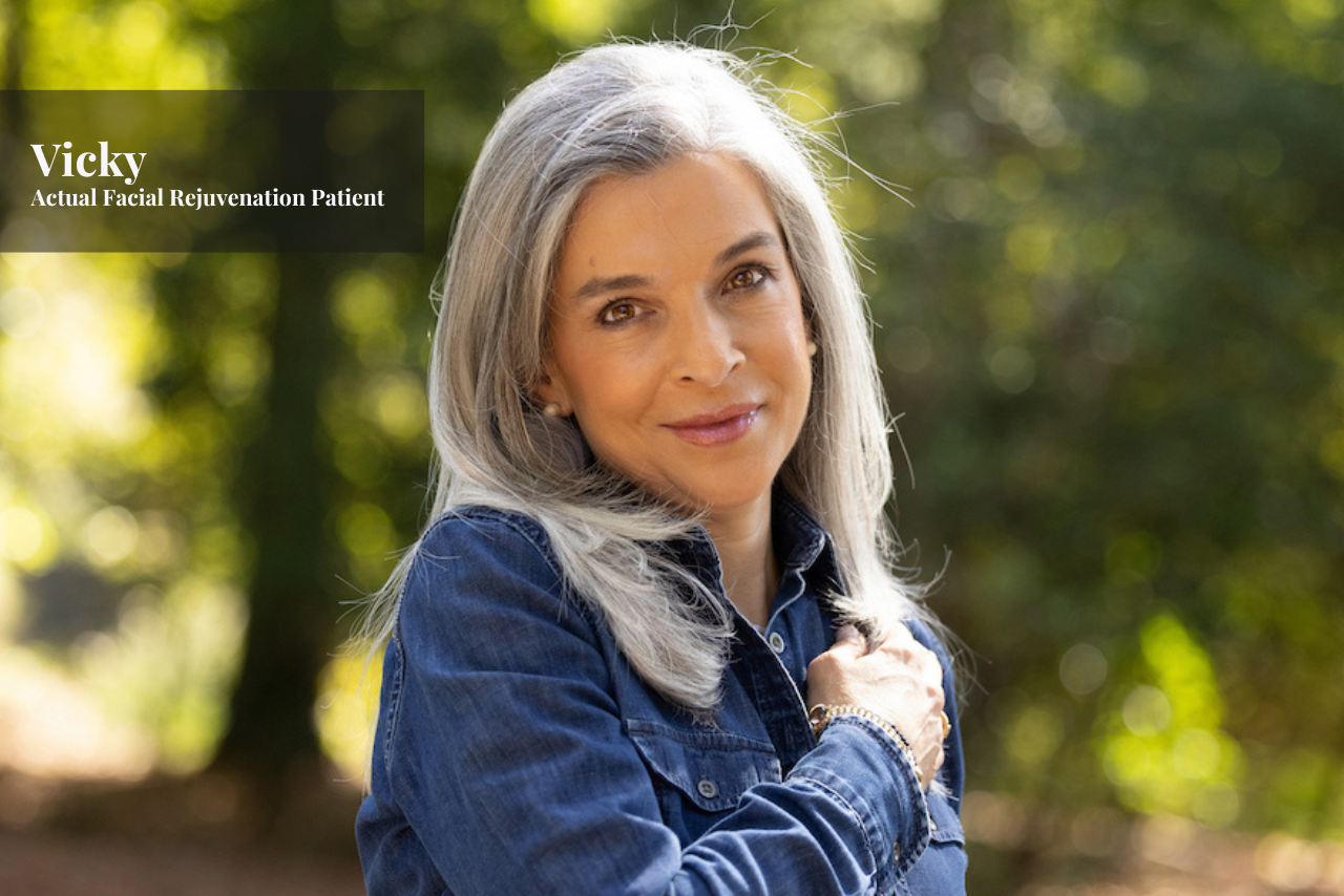 A smiling woman with long gray hair wearing a denim jacket standing outdoors with a blurred background of green trees.