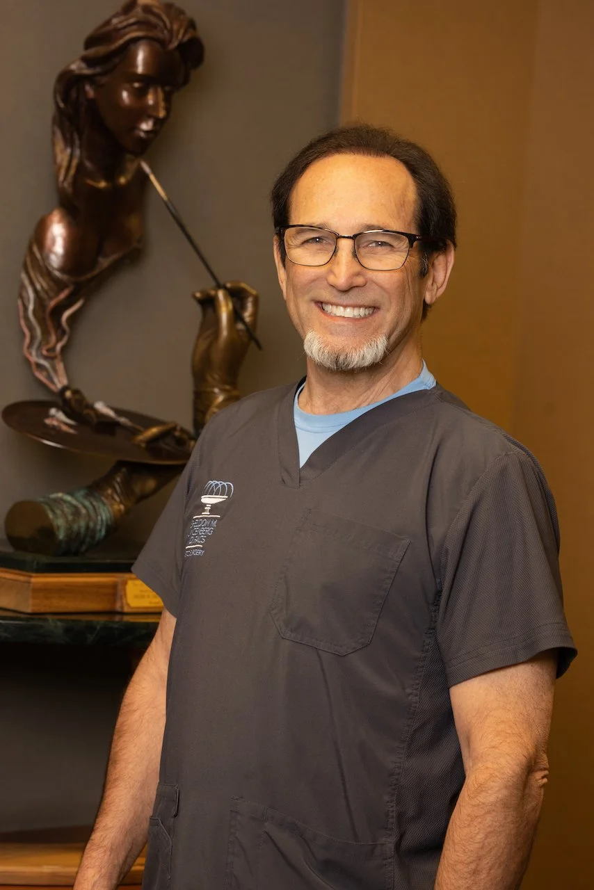 A smiling middle-aged man with glasses and a beard wearing medical scrubs, standing in front of a bronze sculpture of a woman holding a paintbrush.