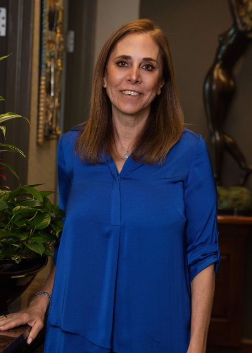 A woman with shoulder-length brown hair wearing a blue blouse standing indoors with plants and artwork in the background.
