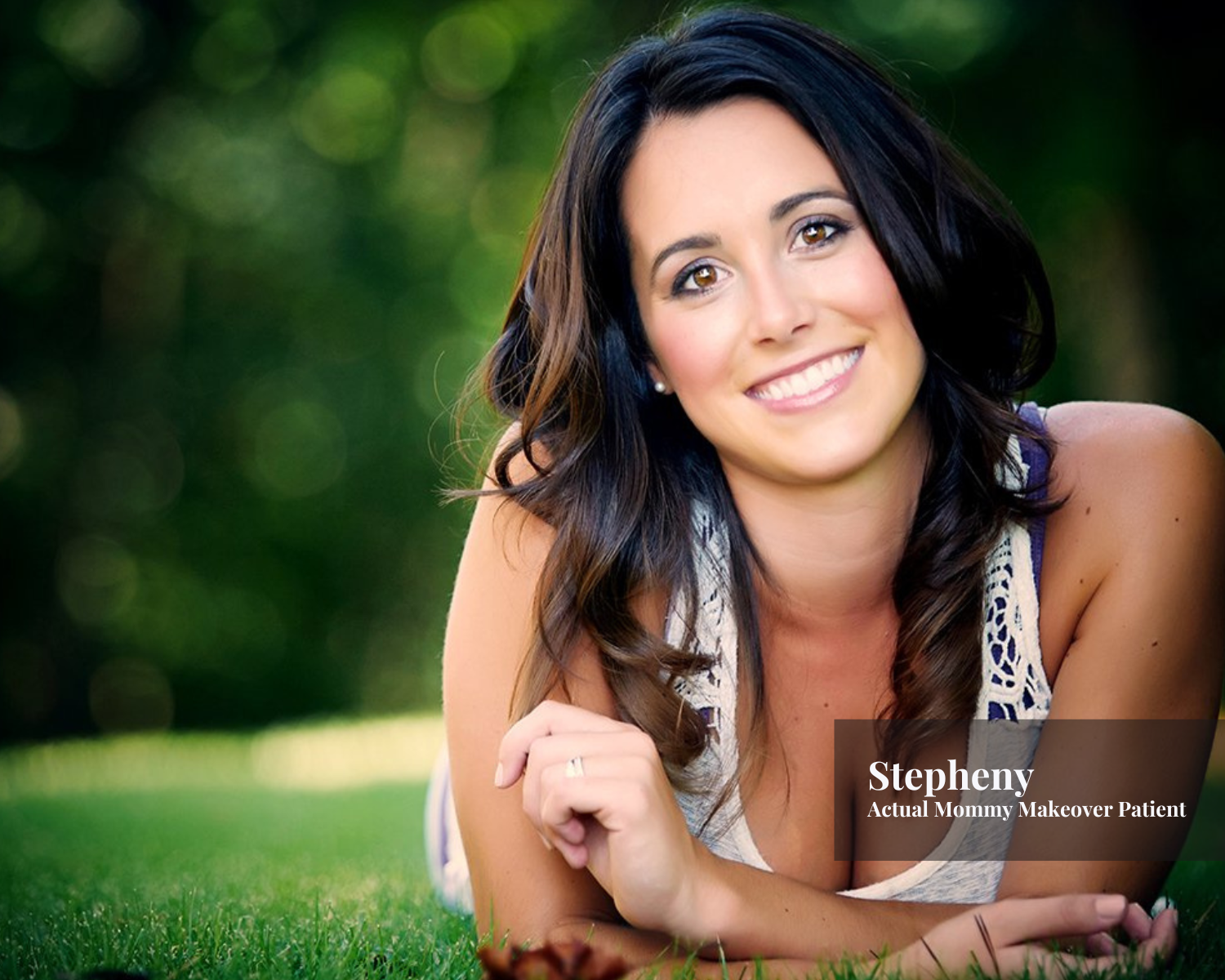 A woman with long dark hair and light makeup lying on the grass outdoors, smiling at the camera, with a blurred green background, labeled 'Stephany, Actual Mommy Makeover Patient'.