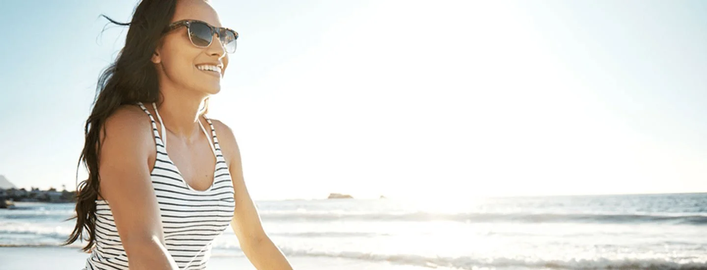 A woman with long dark hair wearing sunglasses and a striped tank top, smiling at the beach during the daytime.