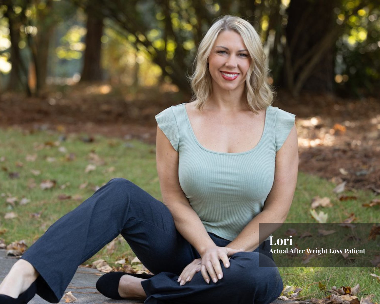A smiling woman with blonde hair sitting on the grass in a park, wearing a light green top and dark blue pants, with fall leaves around her, during daytime.
