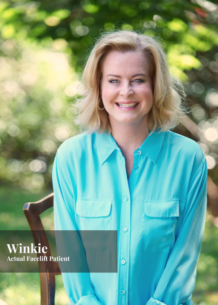 A woman with blonde hair smiling, sitting outdoors with blurred green trees in the background, wearing a light blue button-up shirt.