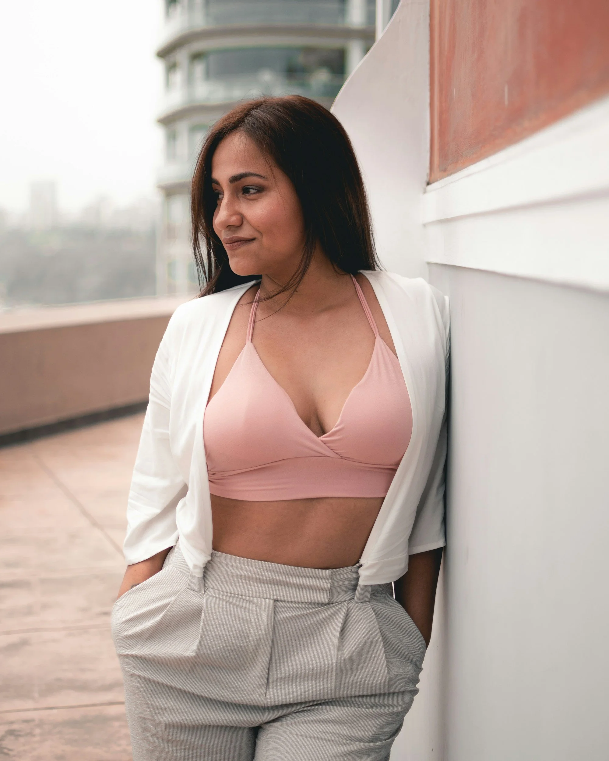 A woman with long dark hair and light skin, wearing a pink top, white blazer, and striped pants, standing outside near a white wall and a building with a rounded balcony in the background.