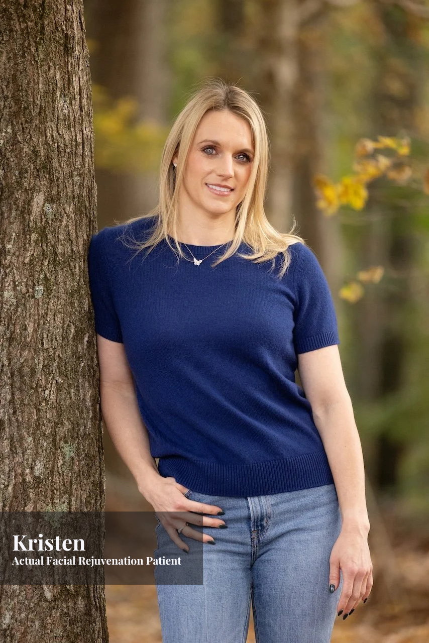 A woman in a blue sweater and jeans leaning against a tree in a forested area with autumn foliage, smiling at the camera.