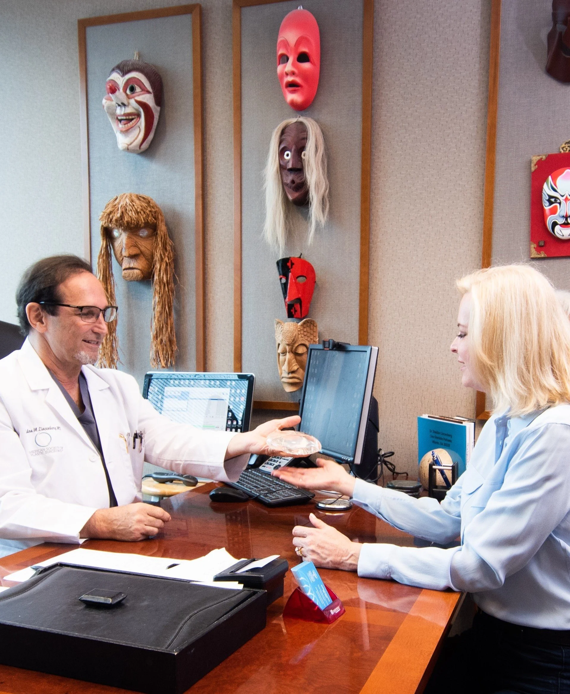 A woman and a man wearing white lab coats are seated at a wooden table in a medical office, engaging in a consultation. The woman has blonde hair and is holding the man's hand, while the man is smiling. Behind them, a wall displays several theatrical masks, including a smiling mask, a sad mask, and others. The scene appears professional and friendly.