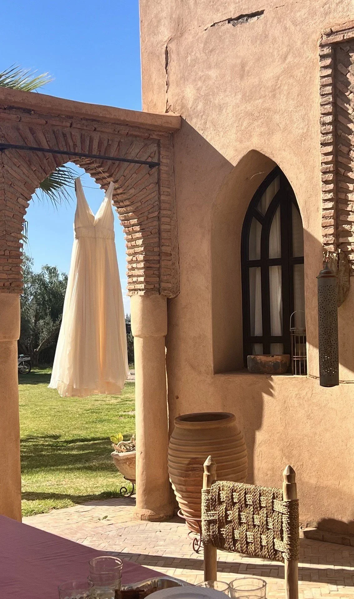 A courtyard scene with beige stucco walls, an arched window with black frame, a large beige ceramic pot, a woven chair, and a white dress hanging on a clothesline, with green grass and trees in the background under a clear blue sky.