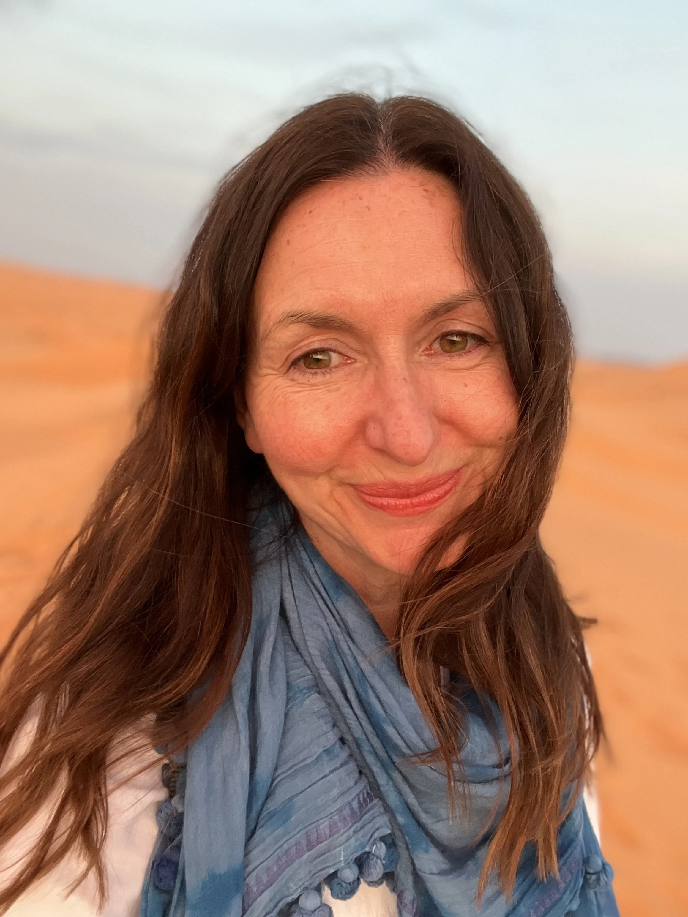 Close-up of a woman with long brown hair smiling, wearing a blue scarf, and standing in front of a blurred background with warm colors.