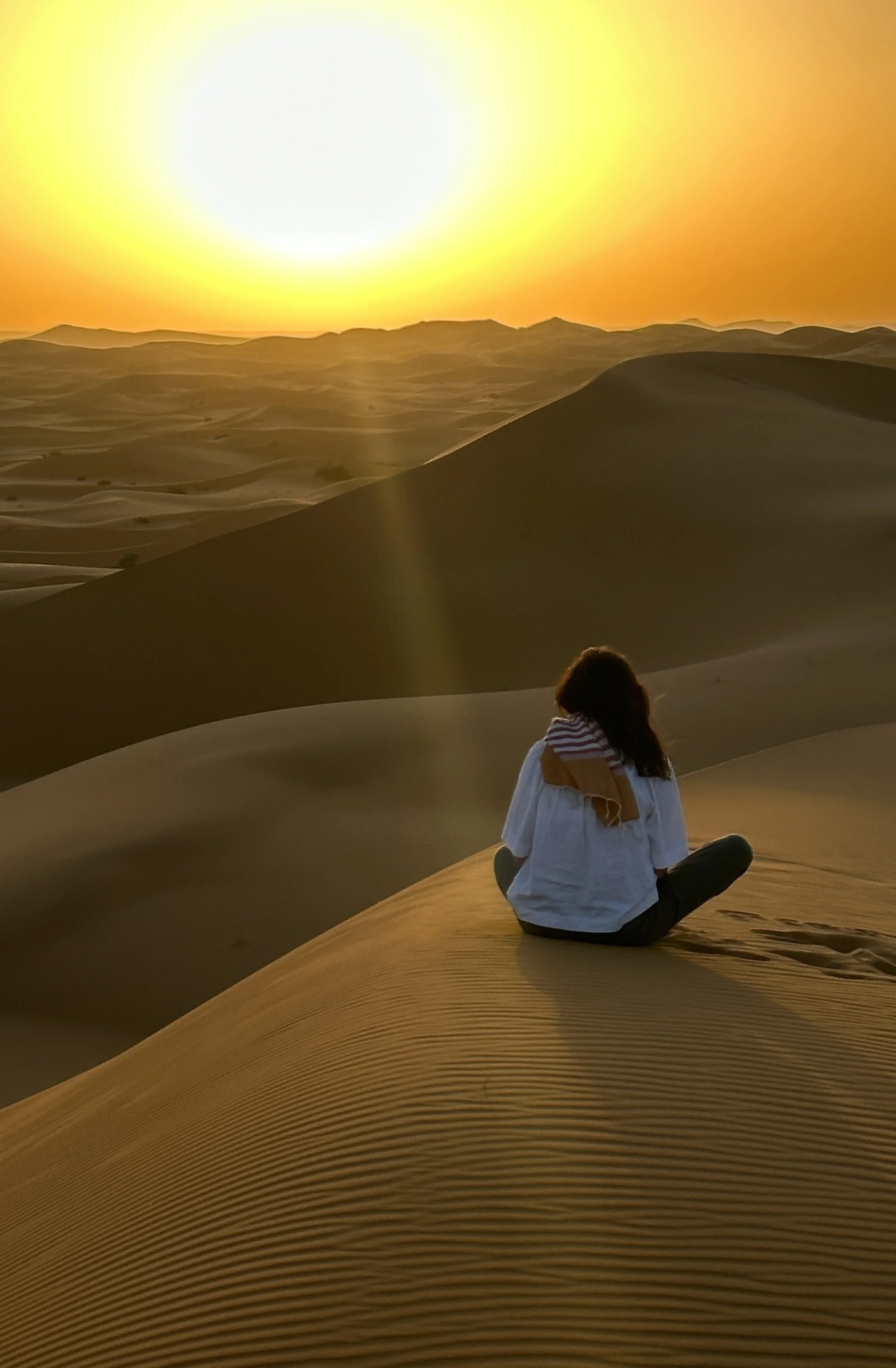 A person sitting cross-legged on sand dunes during sunset or sunrise in a desert landscape, with a woman wearing a white top, jeans, and a striped towel over her shoulders, facing away from the camera.