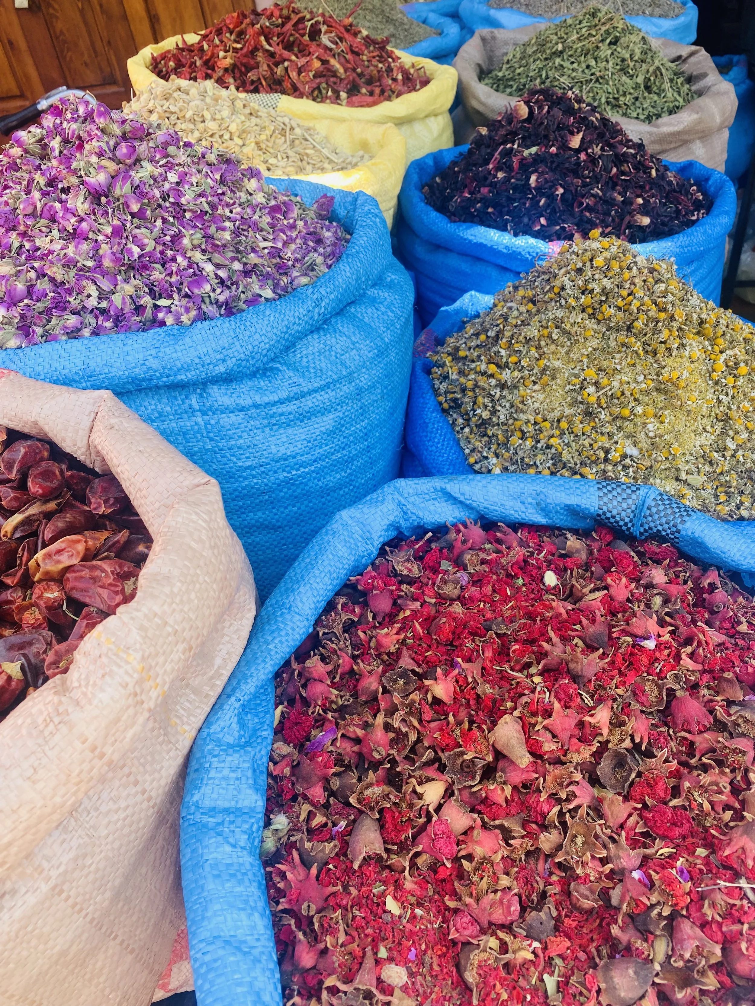 Large sacks of dried flowers and herbs, including rose petals, chamomile, and other aromatic plants, displayed at a market stall.