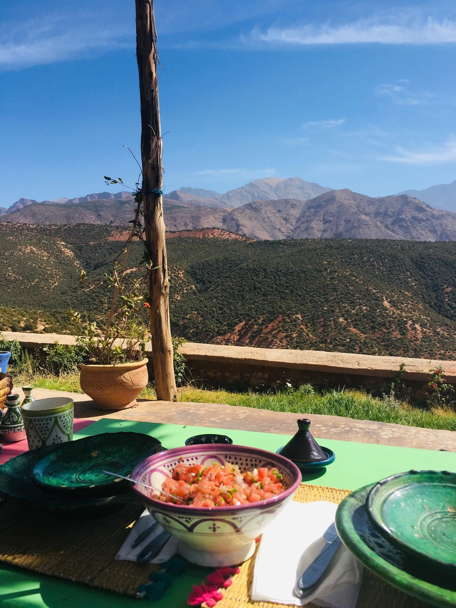 A table set with bowls of food, including a bowl of chopped salad, and surrounded by colorful dishes and utensils, with a scenic mountain view in the background under a blue sky.