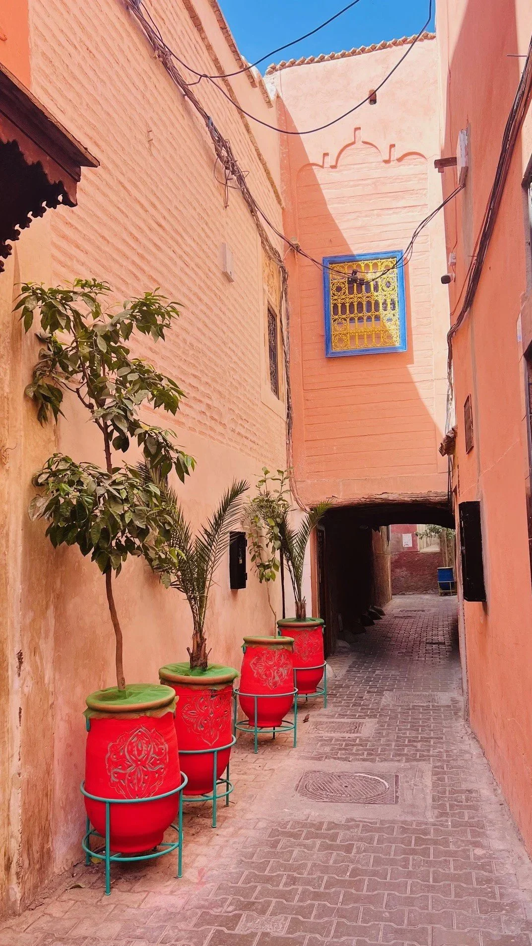 Narrow alley with peach-colored walls, four red pots with plants, underpass at the end, blue sky overhead.
