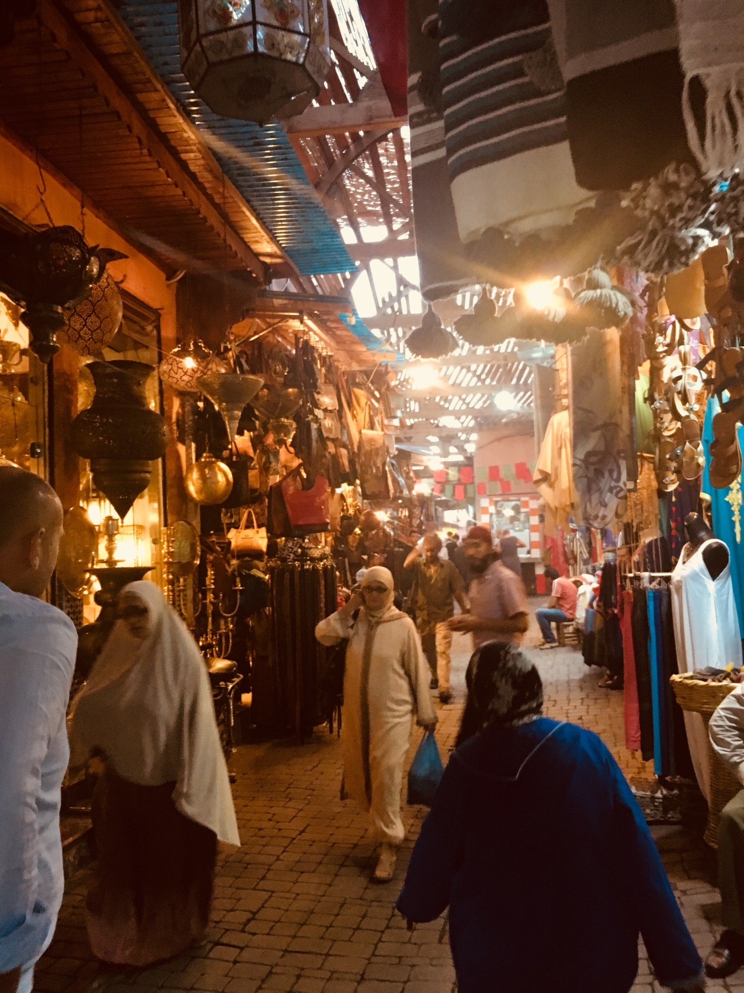 A bustling, indoor market with various hanging lamps, clothing, and decorative items. People are walking and shopping among booths and displays under a roof with wooden beams and banners hanging.