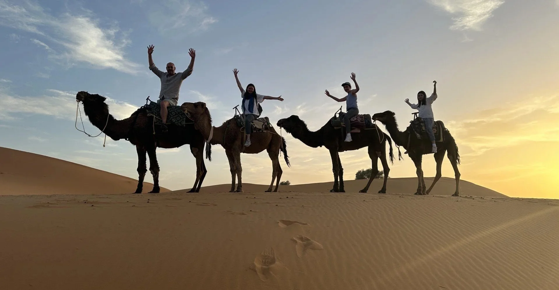 Four people riding camels in a desert at sunset, with footprints in the sand visible in the foreground.