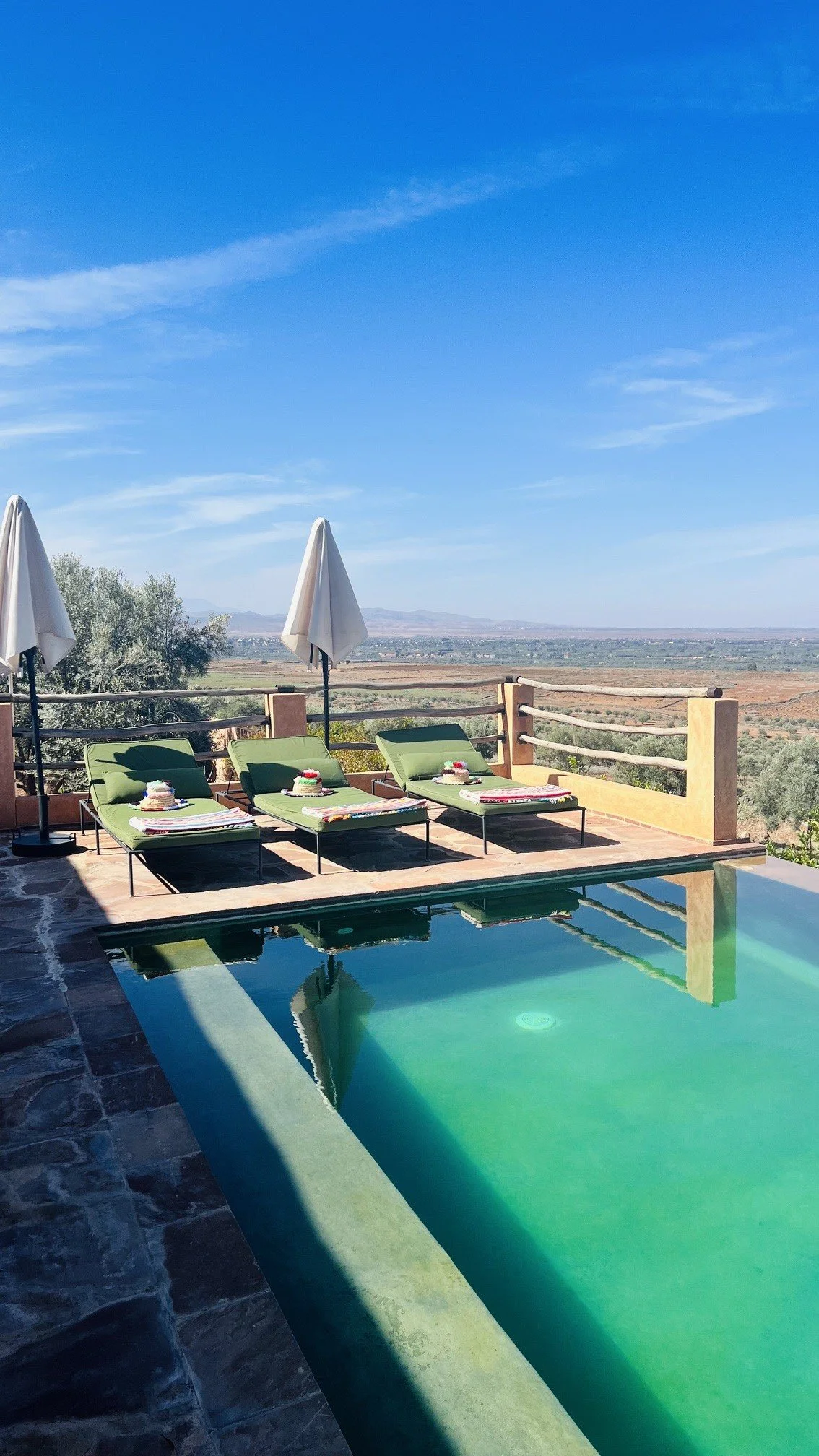 Swimming pool with three green lounge chairs and umbrellas, overlooking a scenic landscape with mountains in the distance under a clear blue sky.