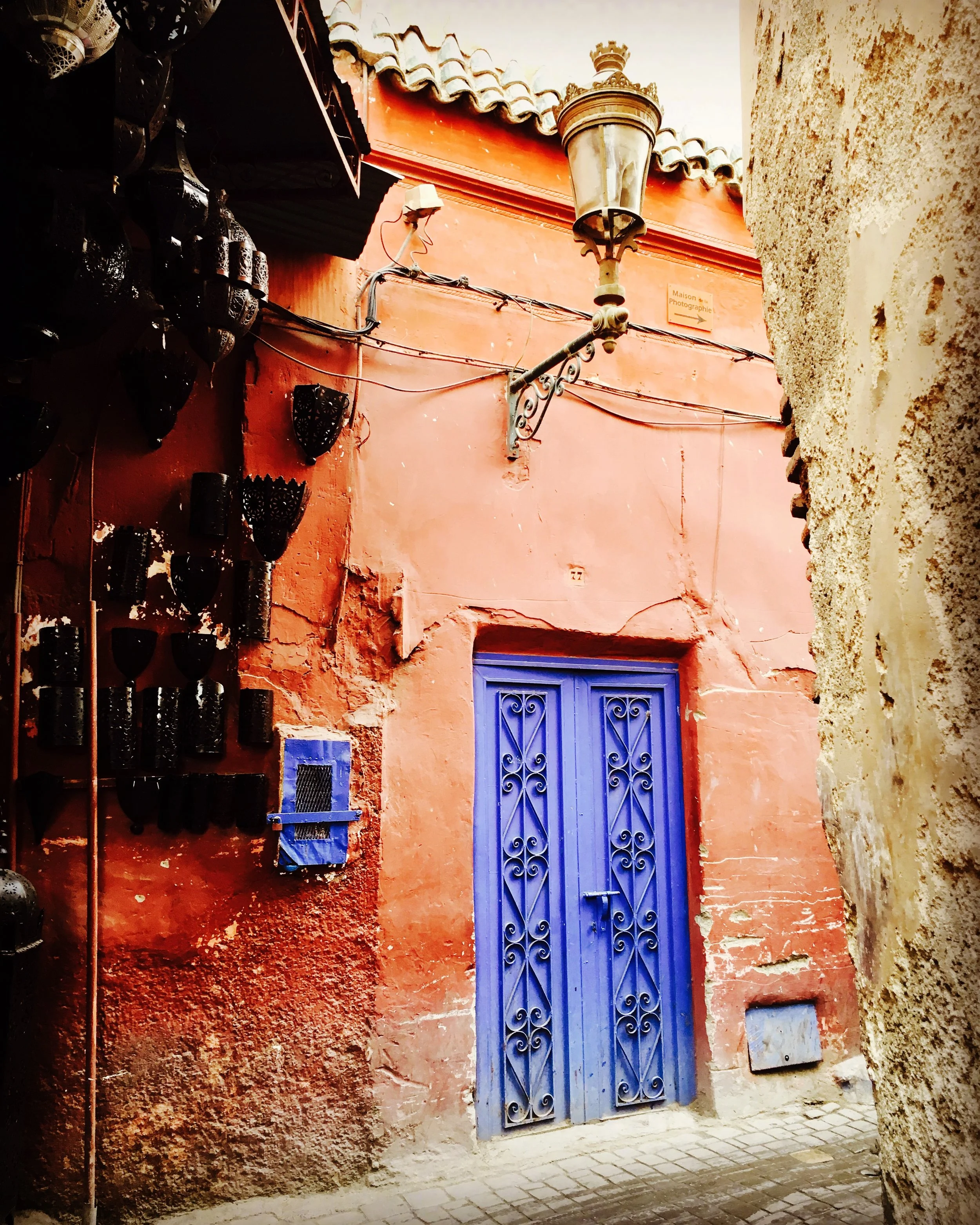A narrow alleyway with a bright blue ornate double door set in a weathered red stucco wall, flanked by a rough stone wall on the right. The alley has cobblestone pavement and features a vintage street lamp, metal wire, and hanging black decorative lanterns on the left.