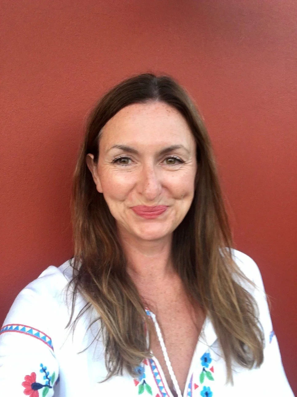A woman with long brown hair and light skin, smiling, wearing a white embroidered blouse, standing against a reddish-brown wall.