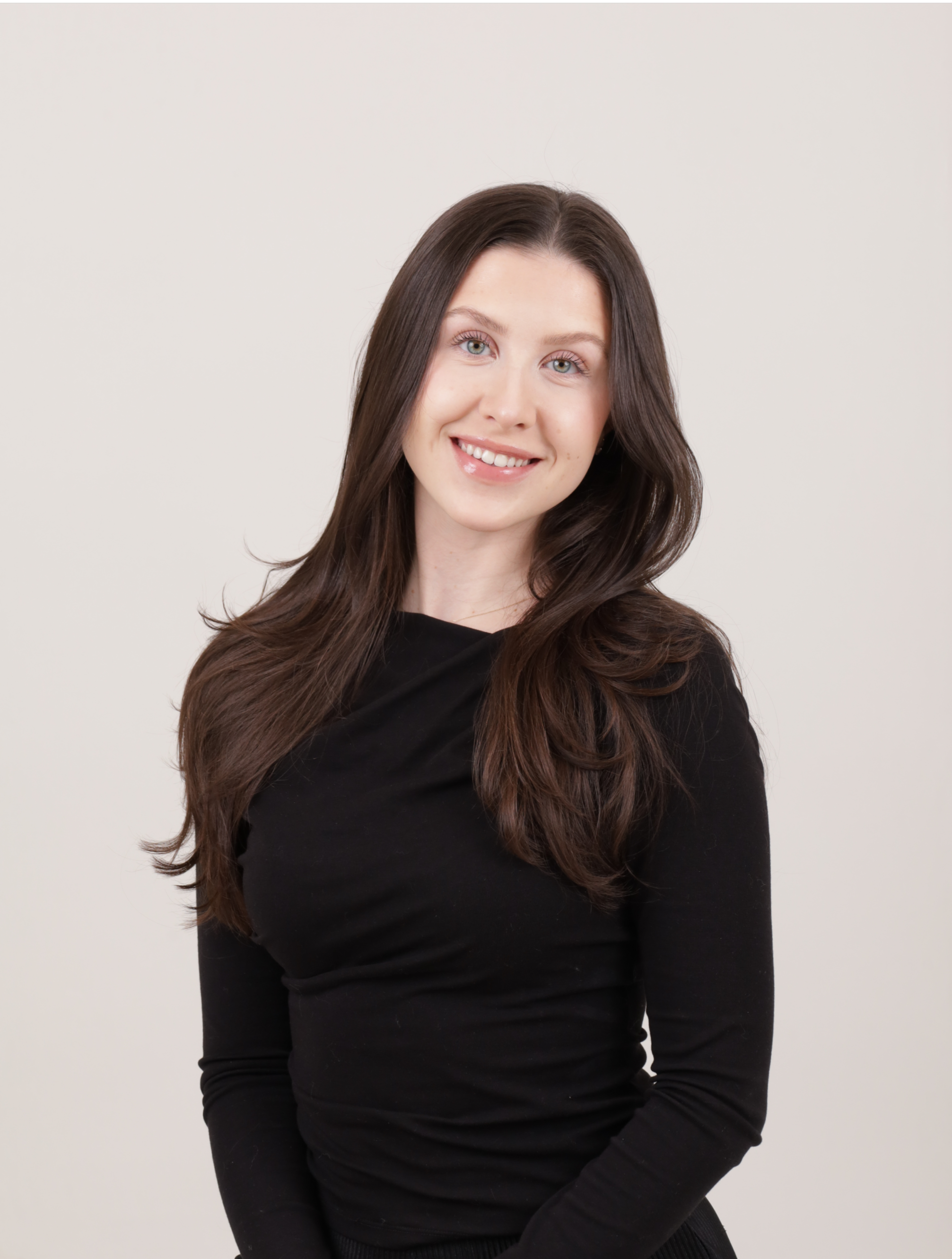 Portrait of a young woman with long, wavy brown hair, smiling, dressed in a black top, standing against a plain light background.