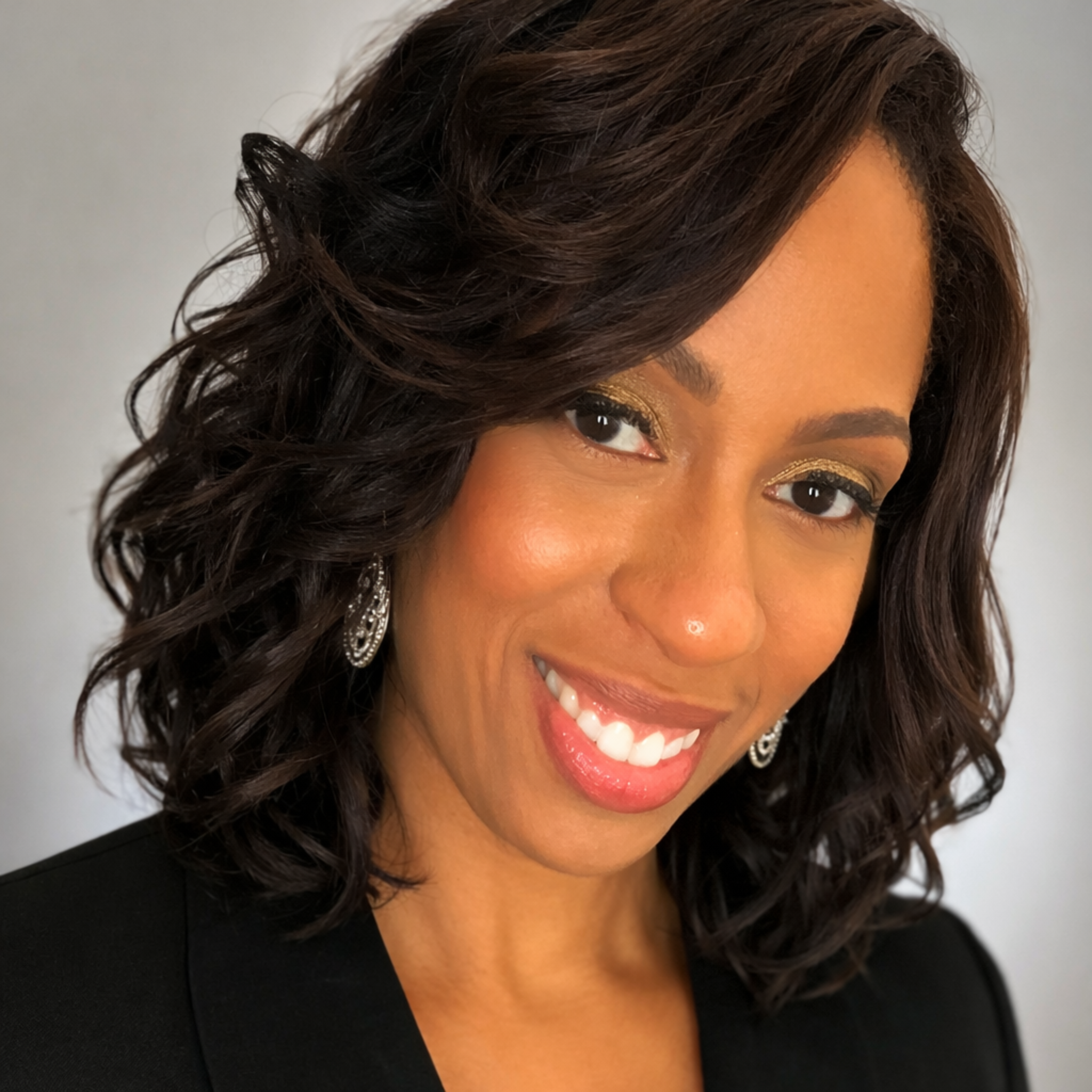 Smiling woman with shoulder-length wavy dark hair, wearing earrings and a black blazer, against a plain light background.